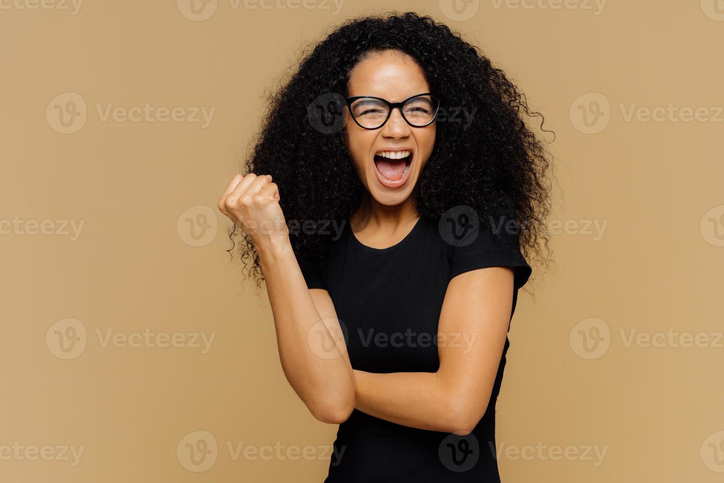 jublande optimistisk kvinna knyter näven, ropar högt, är väldigt känslosam, stöttar sitt favoritfotbollslag, bär avslappnad svart t-shirt, står över brun bakgrund. firande koncept foto