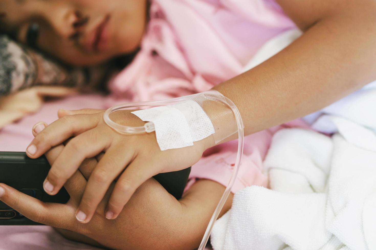barnpatient liggande på sjukhussängen med att spela mobiltelefon och ta emot koksaltlösning. foto