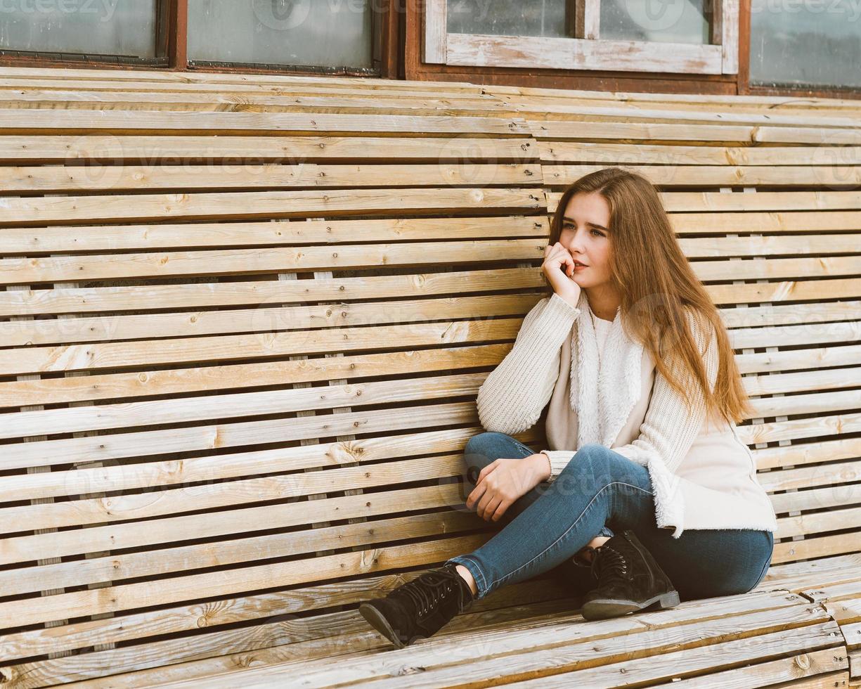 vacker ung flicka med långt brunt hår sitter på träbänk gjord av plankor och vilar, slappnar av och reflekterar. utomhus fotografering med attraktiv kvinna på vintern eller hösten foto