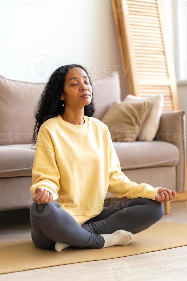 ung afrikansk-amerikansk kvinna sitter i lotus yoga pose på matta i medan mediterar, vertikal foto