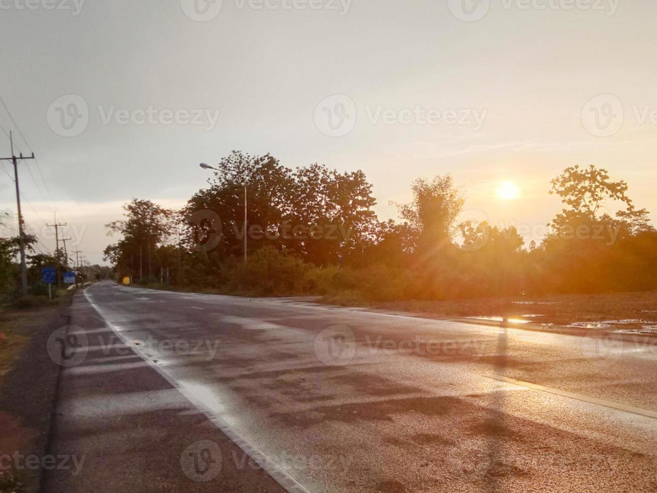 asfalterad väg våt av regn på sidan av vägen bredvid buskträdet, siluettsolnedgången lyser ljus orange färg foto