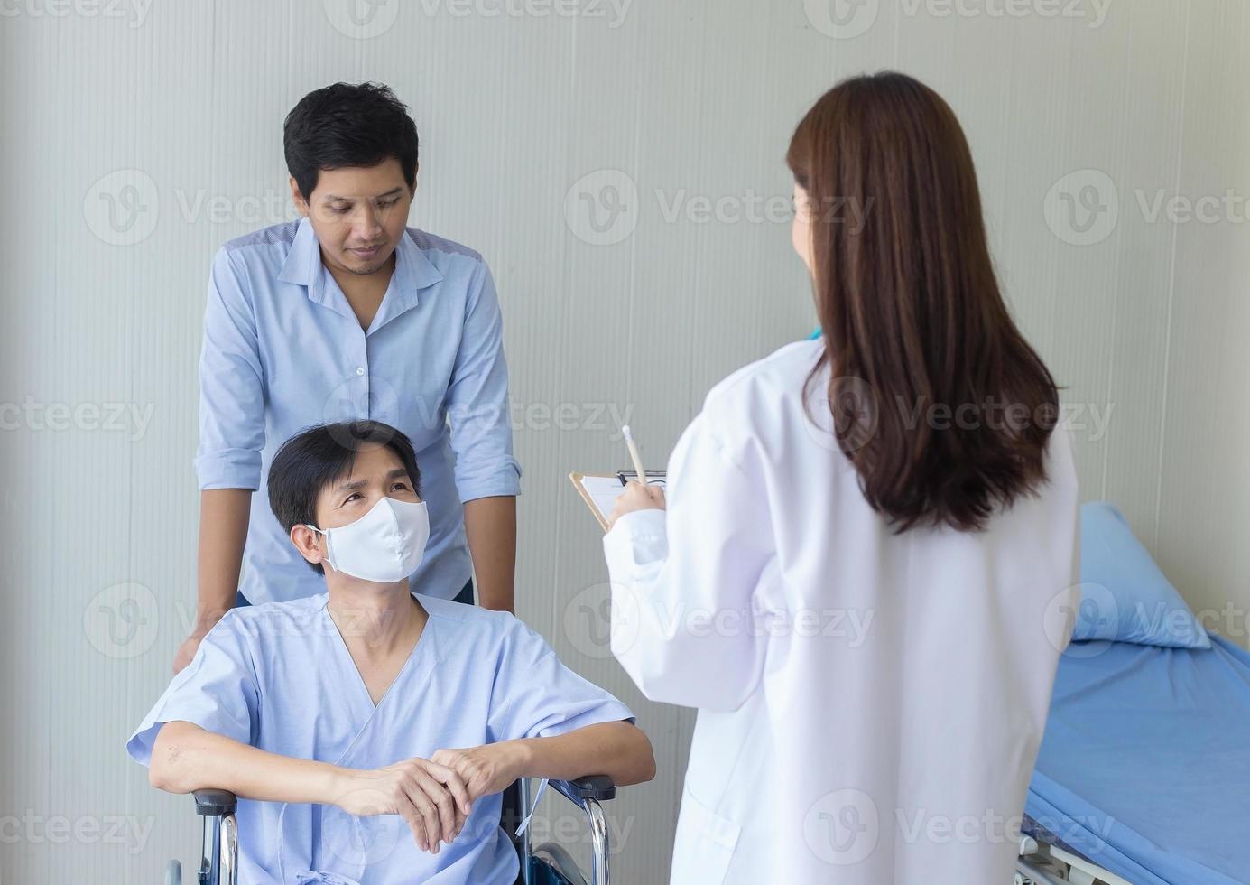 asiatisk kvinna läkare kontrollerar och pratar med en man patient om hans symptom på sjukhus. han bär en kirurgisk mask och sätter sig på rullstolen medan en annan man trycker på den. foto