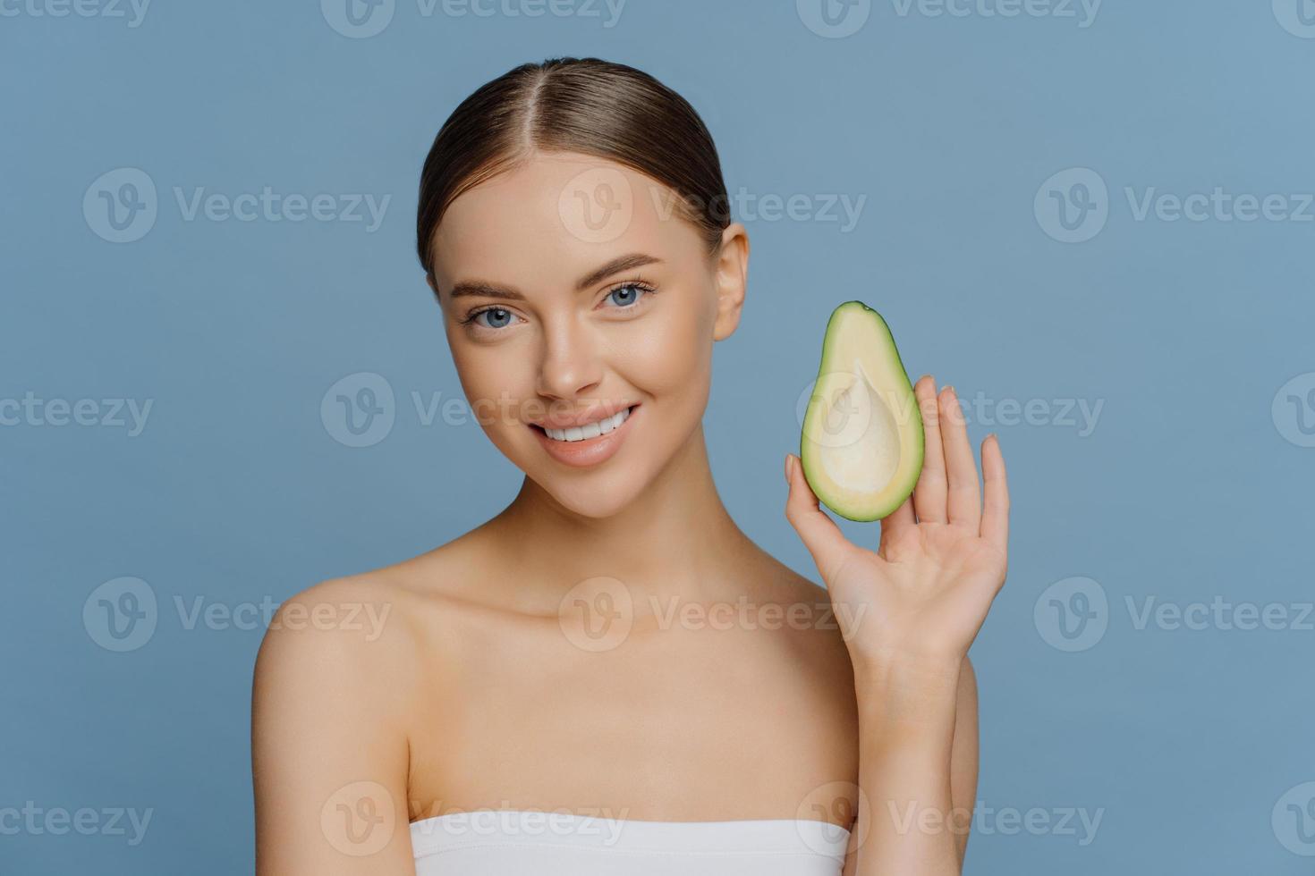 naturlig hudvård koncept. vacker brunett ung kvinna håller hälften av avokado njuter av ekologisk kosmetika njuter av eteriska oljor för skönhetsvård poser insvept i badhandduk mot blå bakgrund foto