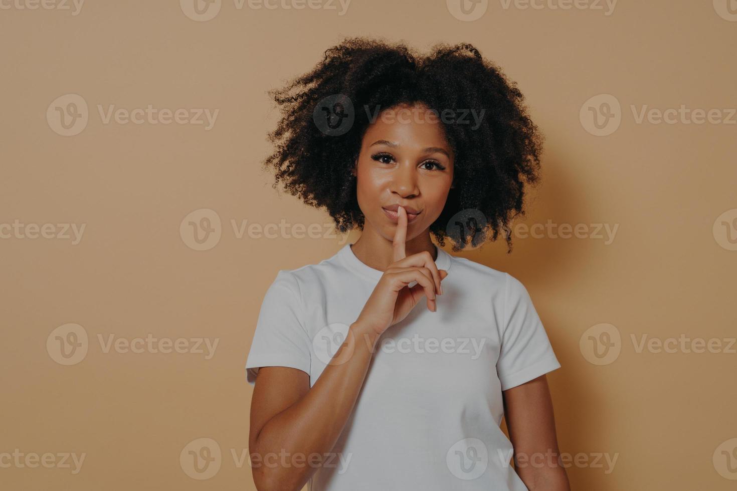 närbild porträtt av afroamerikansk ung våghårig kvinna i vit t-shirt som visar shh-tecken foto