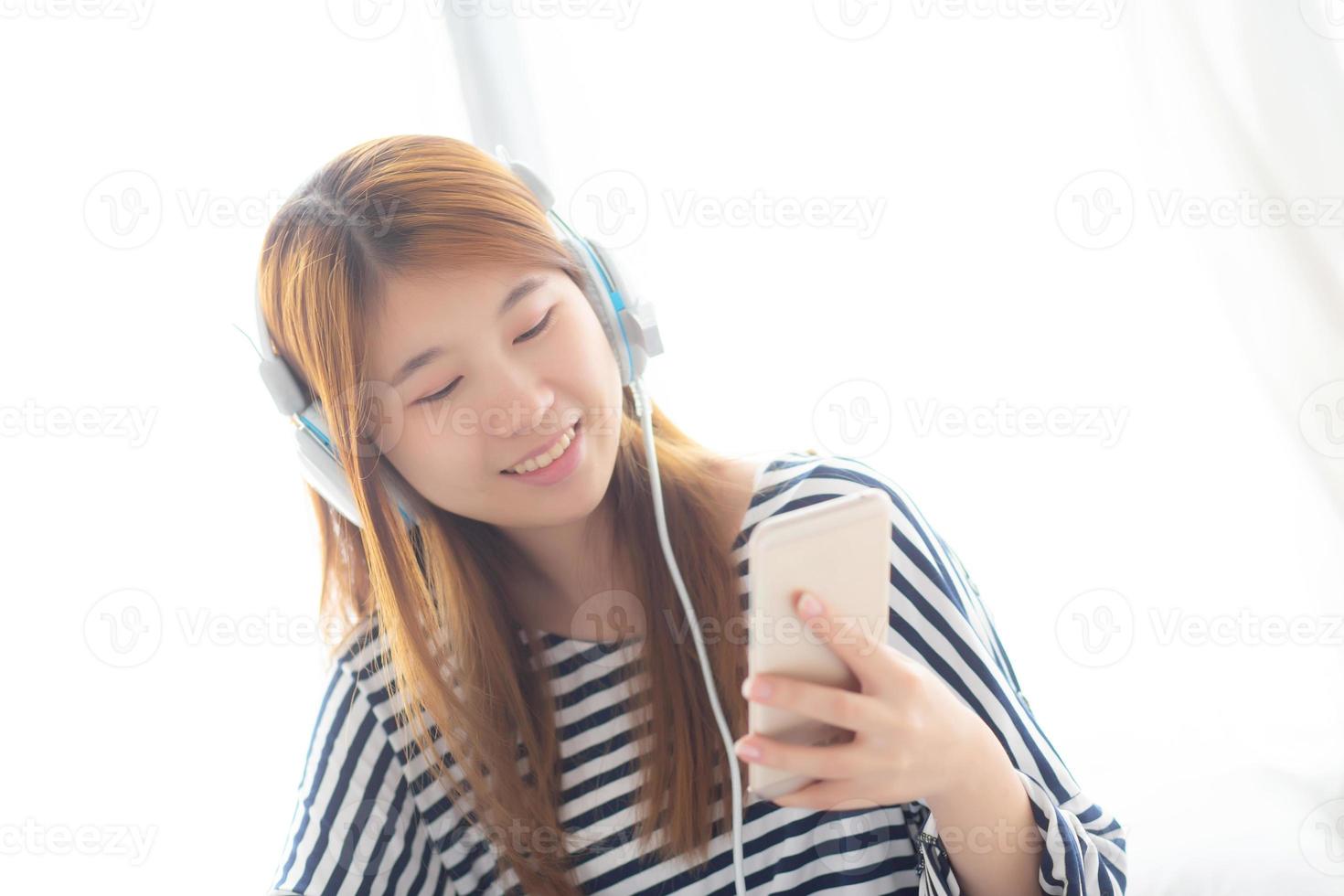 vacker asiatisk ung kvinna njuta av att lyssna på musik med hörlurar och hålla smart mobiltelefon medan du sitter i sovrummet, slappna av tjej med hörlurar, fritid och teknikkoncept. foto