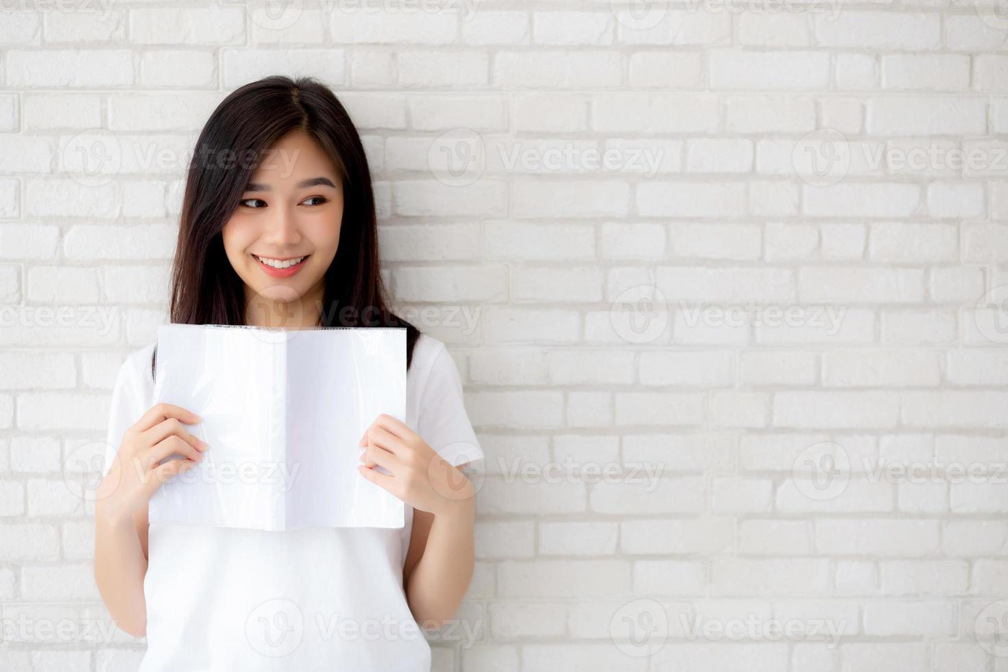 vacker av porträtt ung asiatisk kvinna lycka koppla av stående läsbok på betongcement vit bakgrund hemma, flicka glad studieinnehåll litteratur, utbildning och livsstilskoncept. foto