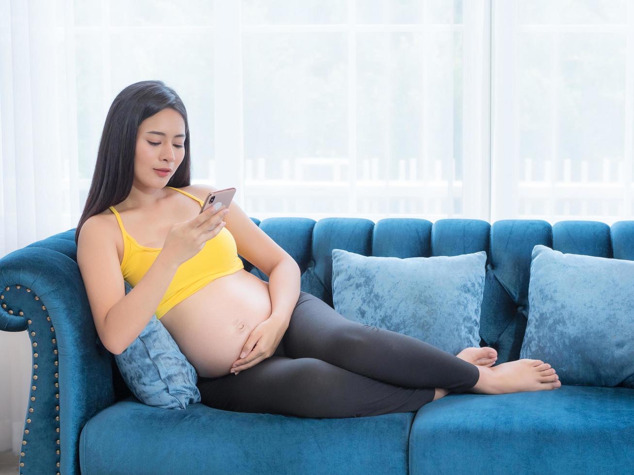 en vacker gravid asiatisk kvinna sitter i soffan och använder en mobiltelefon för att söka information om sitt barn till sin kommande baby foto