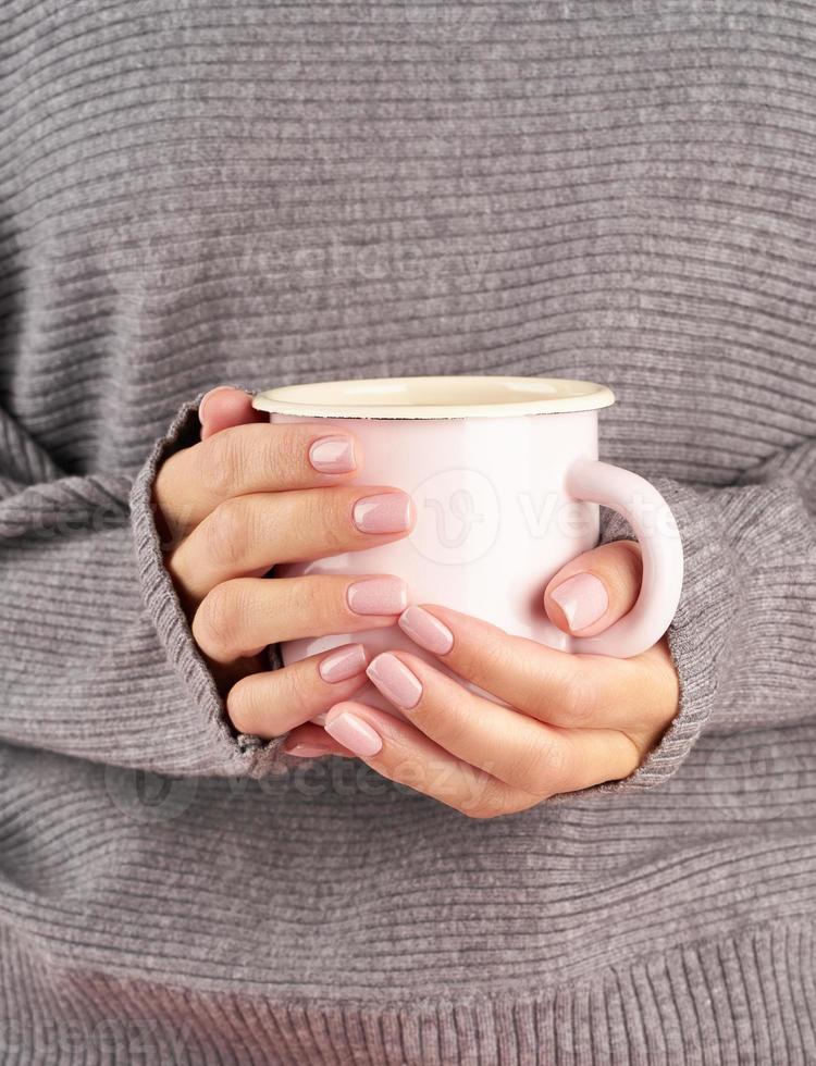 varmt morgonkaffe på jobbet en kall höstmorgon, händer som håller en mugg med en drink, grå tröja, rosa manikyr, närbild, vertikal foto