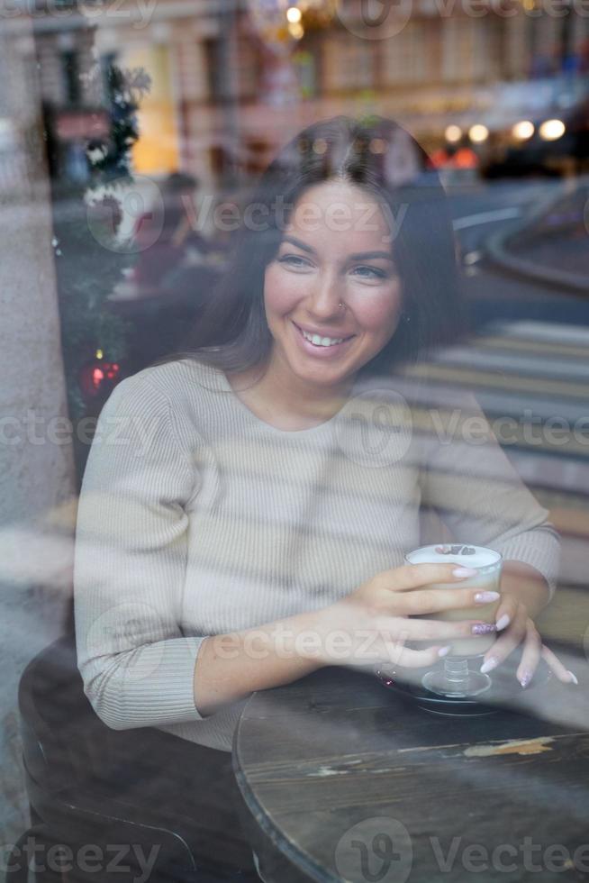 en vacker flicka sitter på ett kafé och tittar eftertänksamt ut genom fönstret. reflektion av staden i fönstret. leende brunett kvinna med långt hår dricker cappuccino, vertikal foto