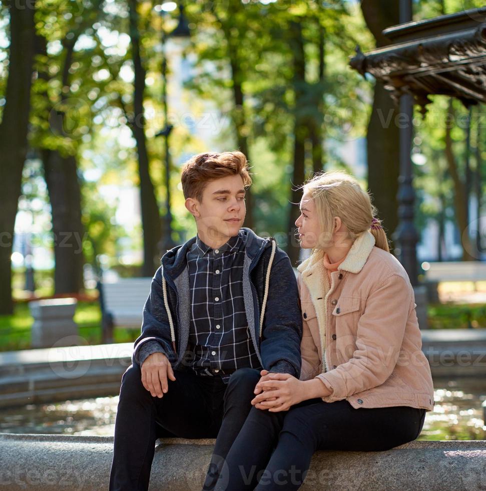 älskare pojke och flicka tittar på varandra, sitter i parken nära fontänen på hösten. flickvän och pojkvän pratar konceptet kärlek och lycka foto