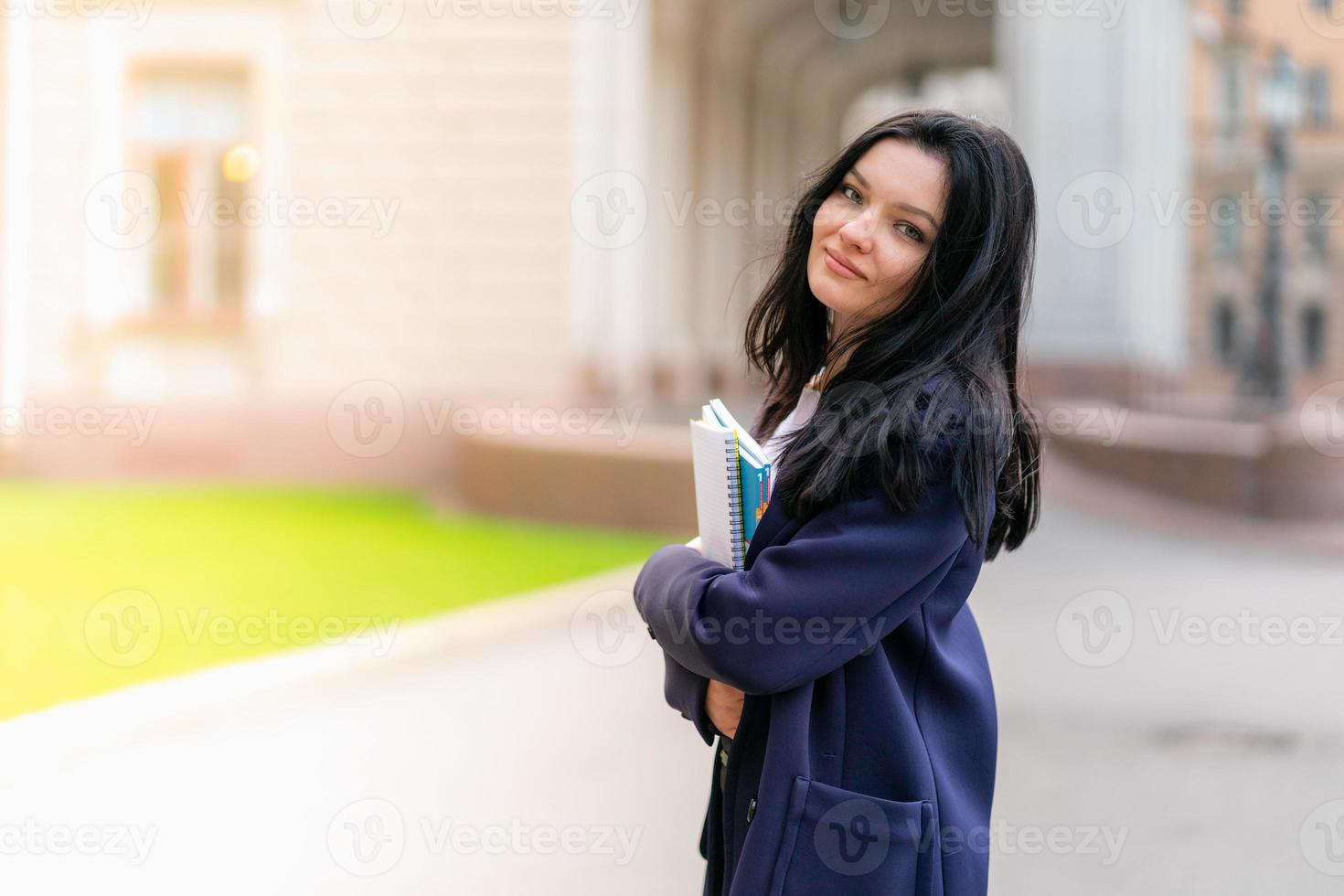 vacker leende flicka brunett student håller anteckningsböcker och läroböcker, står på universitetet på gatan av st. petersburg. en charmig kvinna med långt mörkt hår studerar på kurs, copy space foto