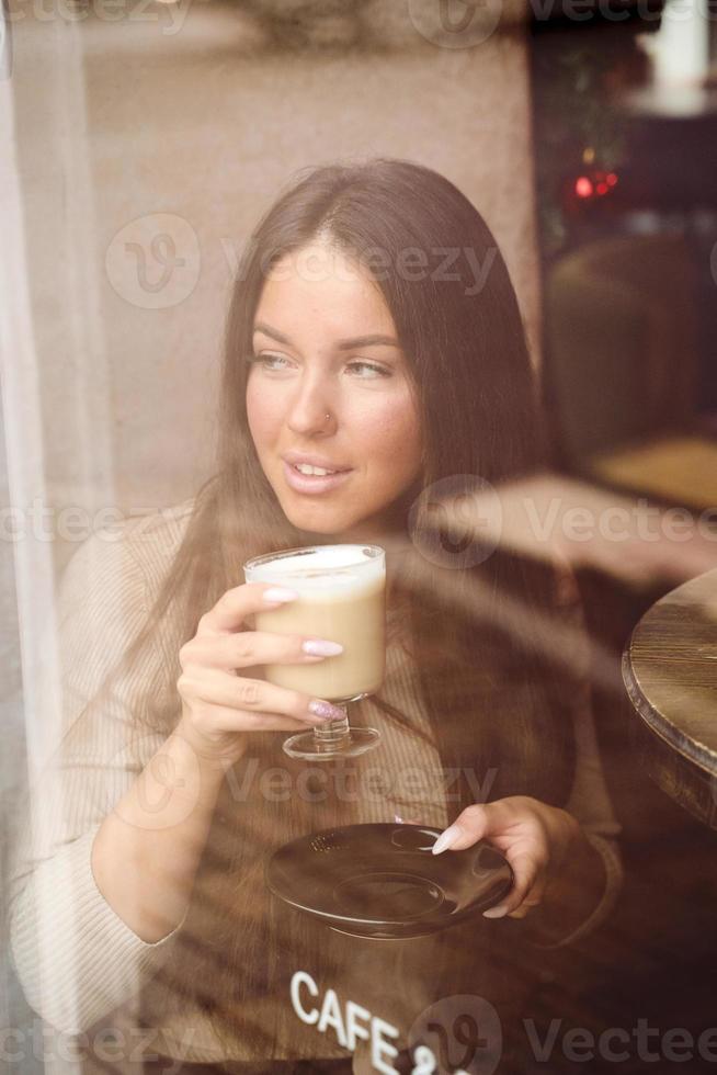 en vacker flicka sitter på café och tittar eftertänksamt ut genom fönstret. reflektion av staden i fönstret. brunett kvinna med långt hår dricker cappuccino, vertikal foto