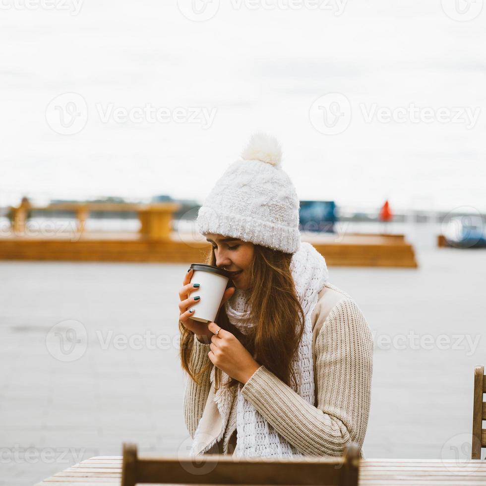 vacker ung flicka dricker kaffe, te från plastmugg i höst, vinter. en kvinna med långt hår i varma kläder som sitter vid vattnet vid Östersjön i hamn, värmd av varm dryck foto