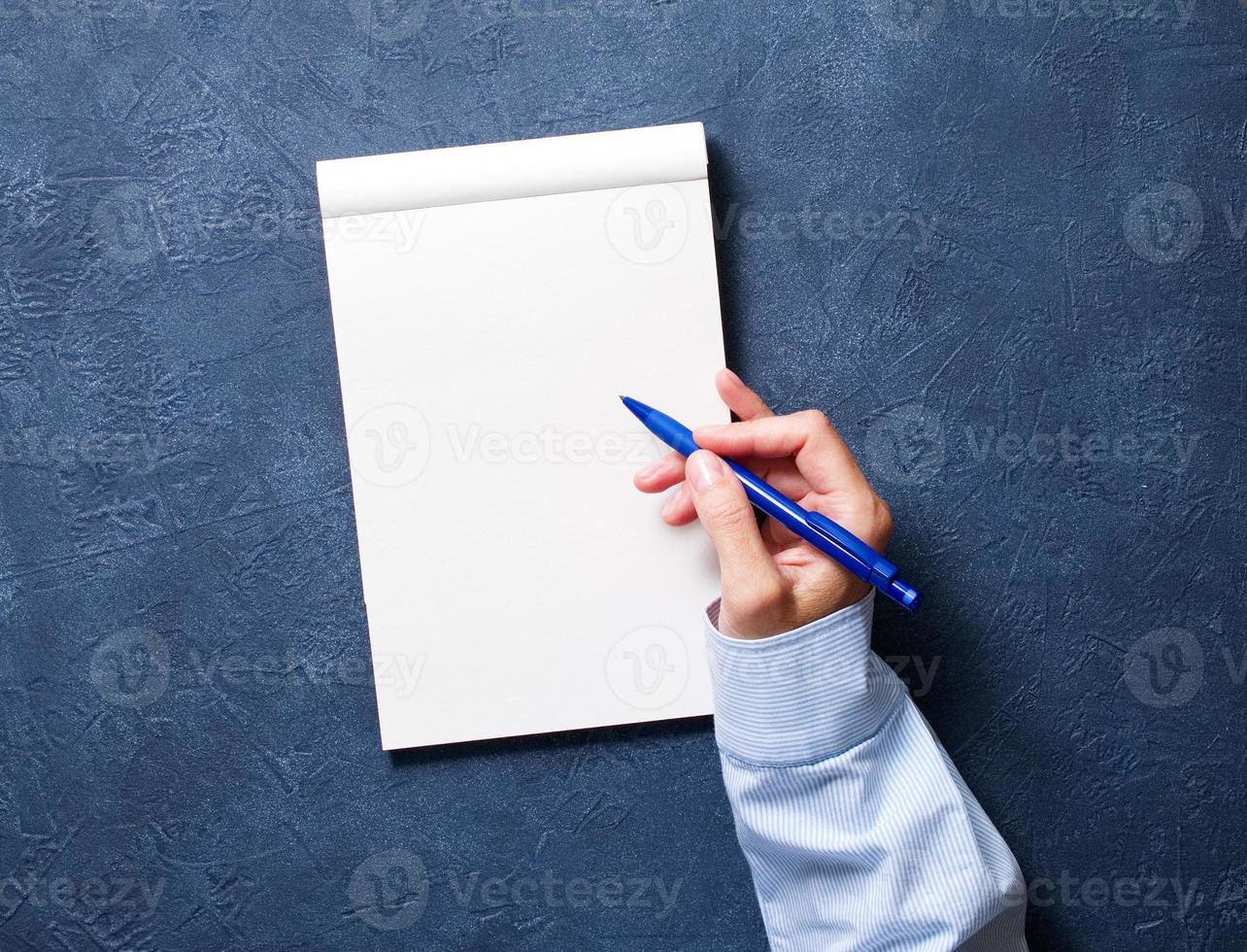 kvinna skriver i en anteckningsbok på mörkblått bord, hand i skjortan med en penna, skissbokteckning foto