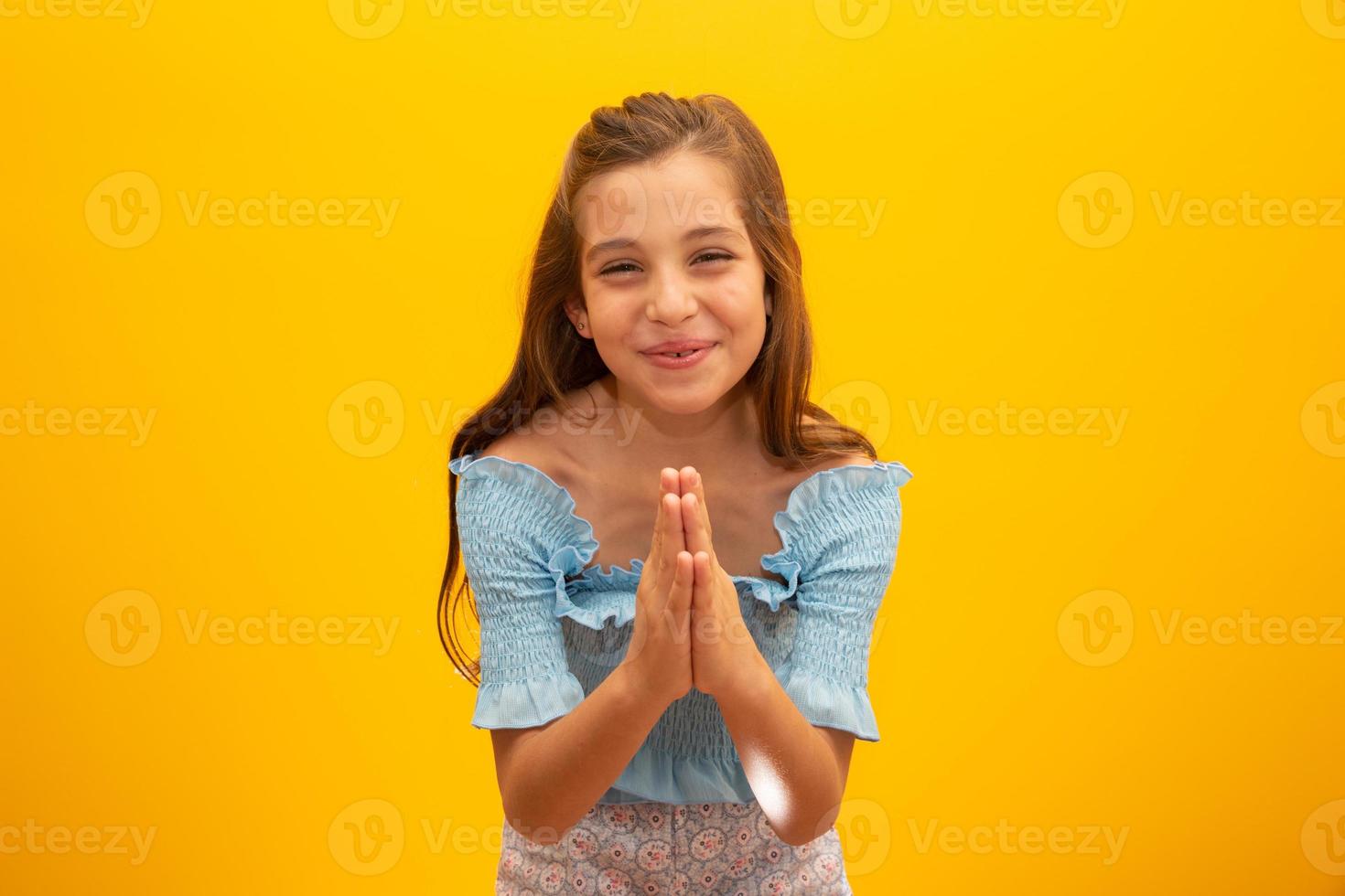 trevlig liten flicka, håller händerna i en bedjande gest, står över isolerad gul bakgrund med kopia utrymme. foto