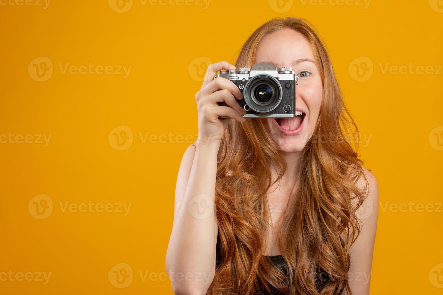 foto av bedårande rödhårig kvinna som bär svarta kläder som håller retro vintagekamera och tar en bild isolerad över gul bakgrund. mock up kopia utrymme. hålla retro vintage fotokamera.