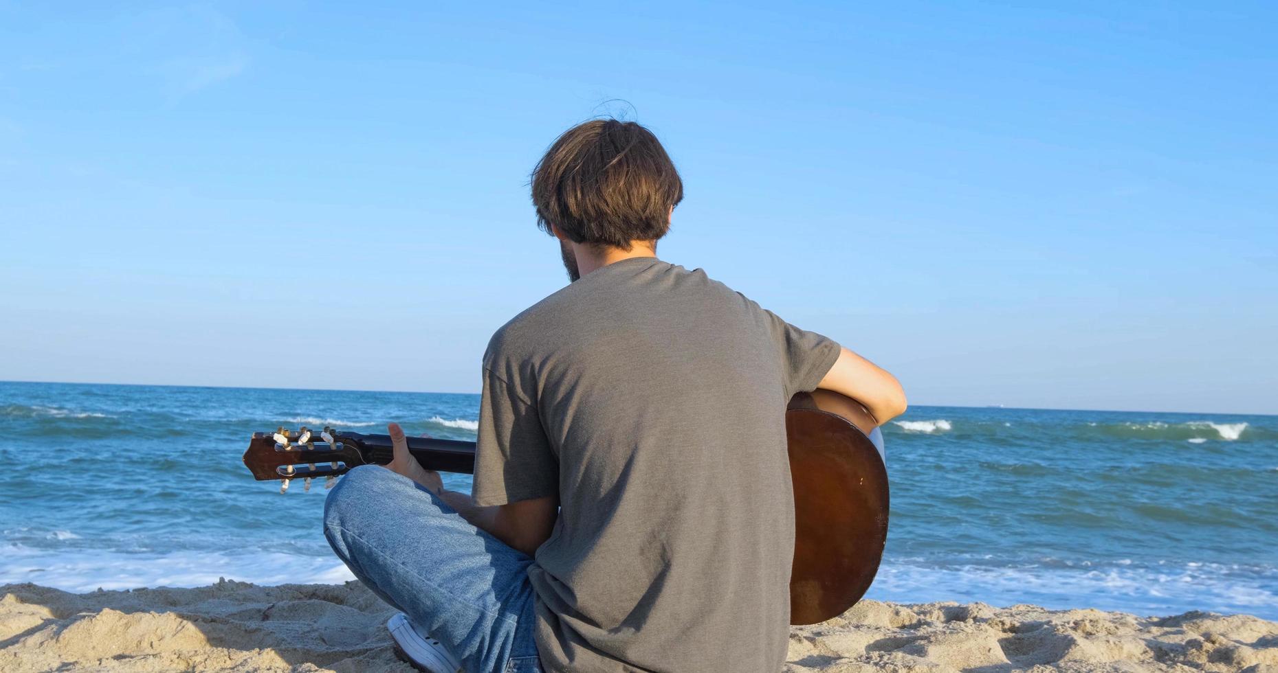ung stilig man spelar i akustisk gitarr på stranden i solig dag, havet eller havet på bakgrunden foto