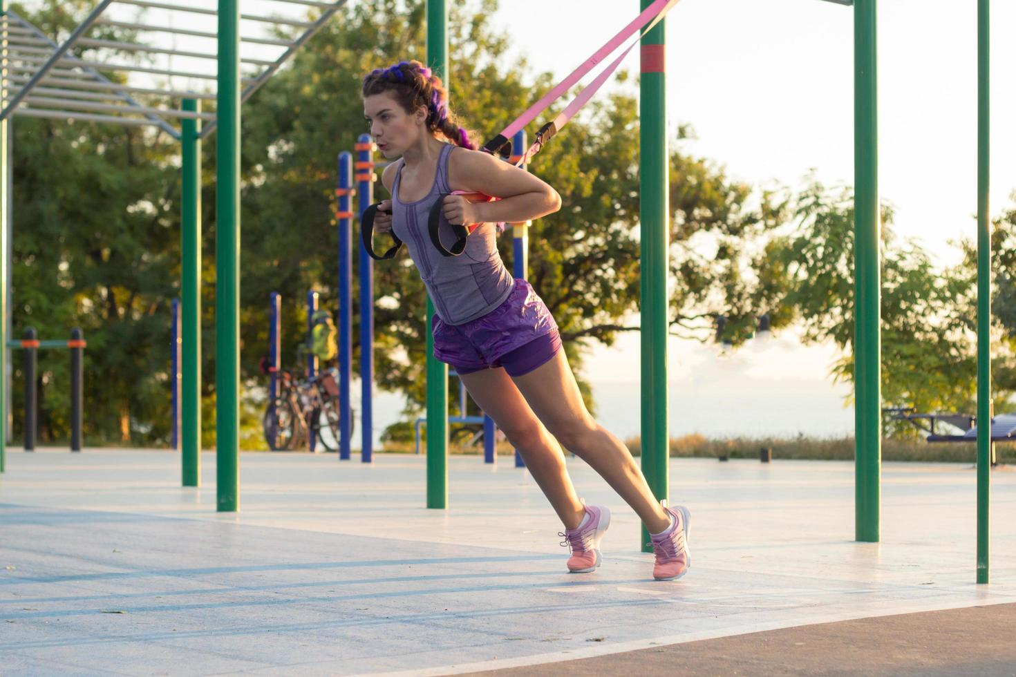 vacker passform kvinna i rosa och lila sportkläder träning på utomhusgym på morgonen, övningar med upphängningsremmar i parken foto