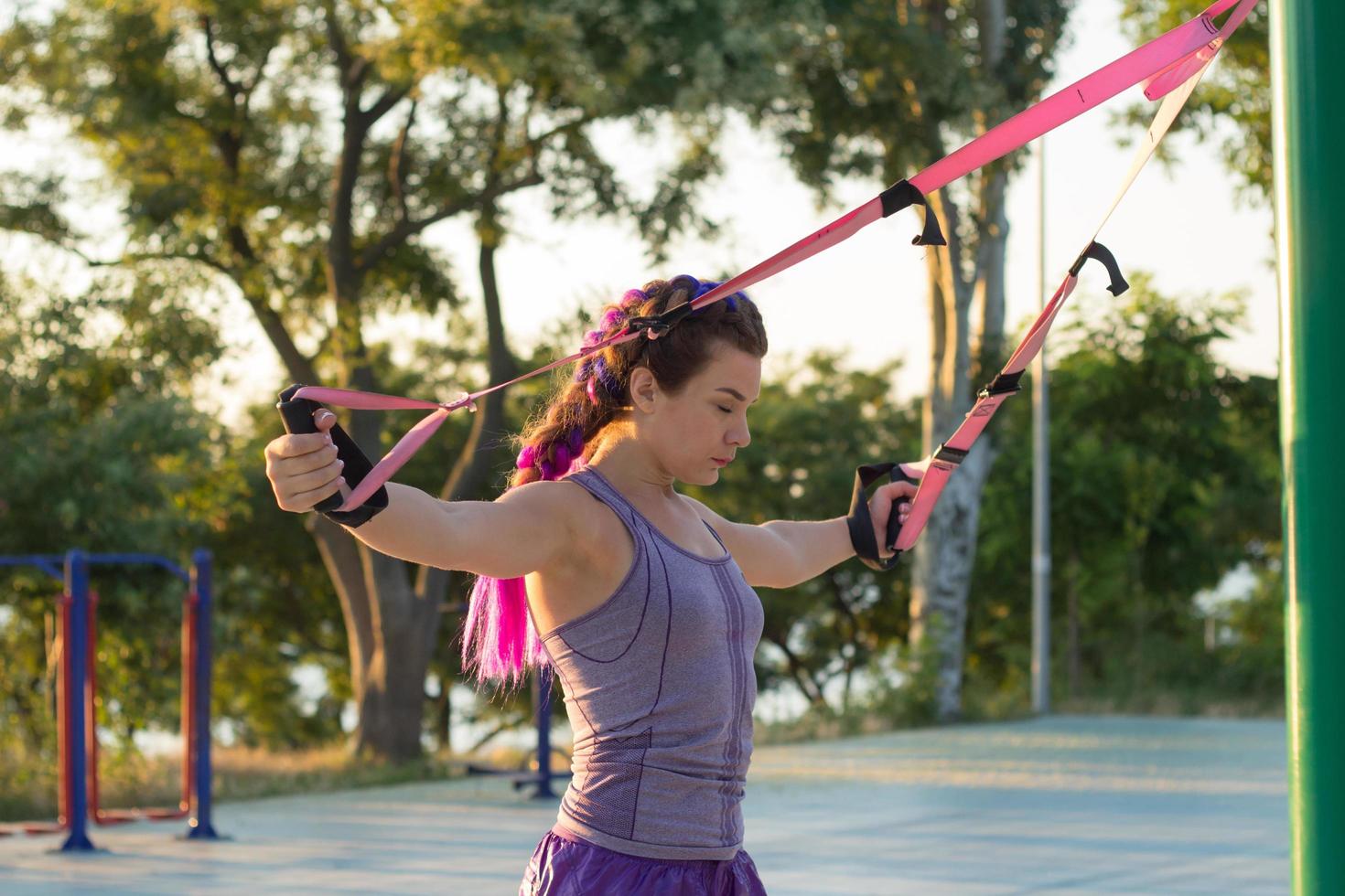 vacker passform kvinna i rosa och lila sportkläder träning på utomhusgym på morgonen, övningar med upphängningsremmar i parken foto