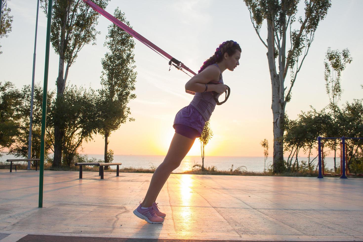 vacker passform kvinna i rosa och lila sportkläder träning på utomhusgym på morgonen, övningar med upphängningsremmar i parken foto