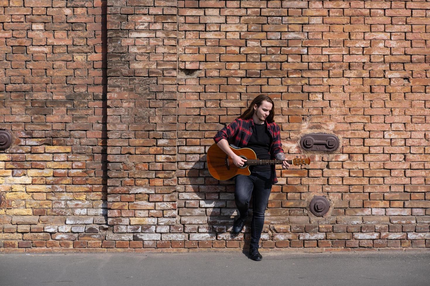 ung man med långt hår och tatuering spelar på akustisk gitarr utomhus på gatan foto