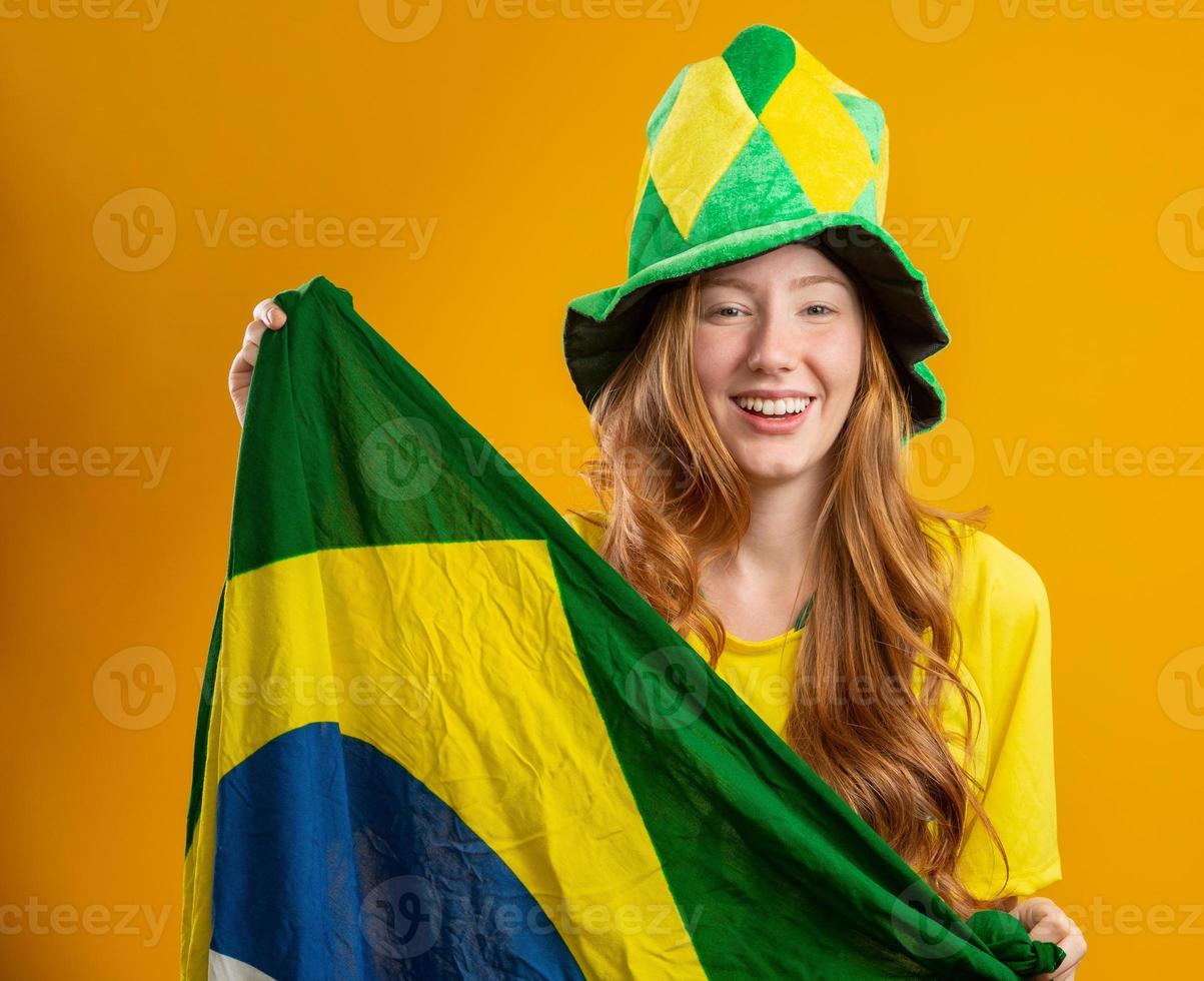 brasiliansk supporter. brasiliansk rödhårig kvinna fan firar på fotboll, fotbollsmatch på gul bakgrund. brasilianska färger. iklädd t-shirt, flagga och fanhatt. foto