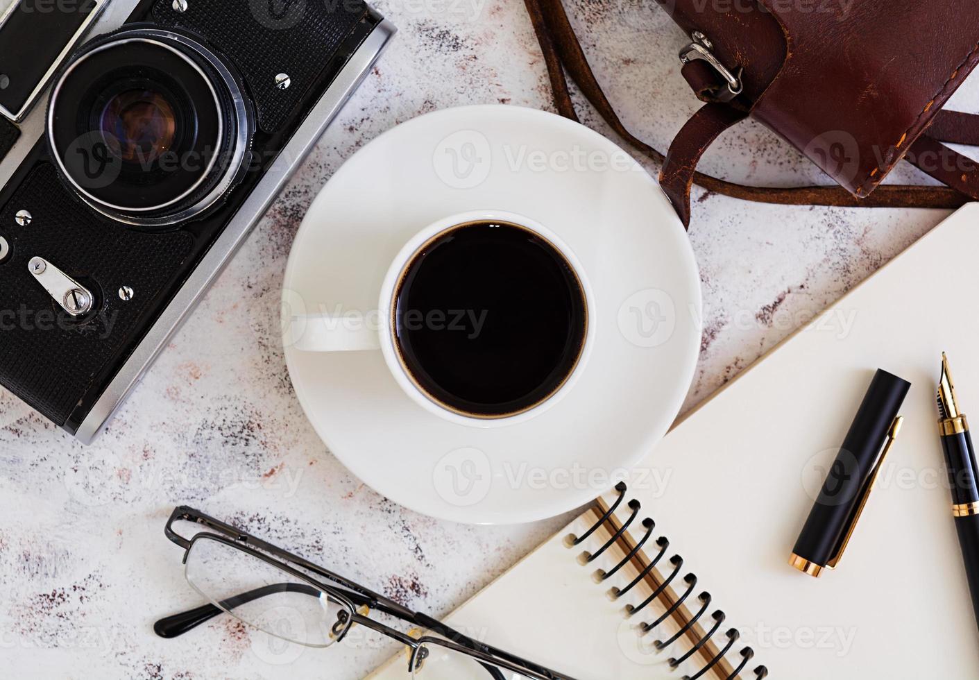 platt låg, ovanifrån kontorsbord skrivbord. skrivbordsarbetsyta med retrokamera, dagbok, penna, glasögon, fodral, kopp kaffe på vit bakgrund. foto