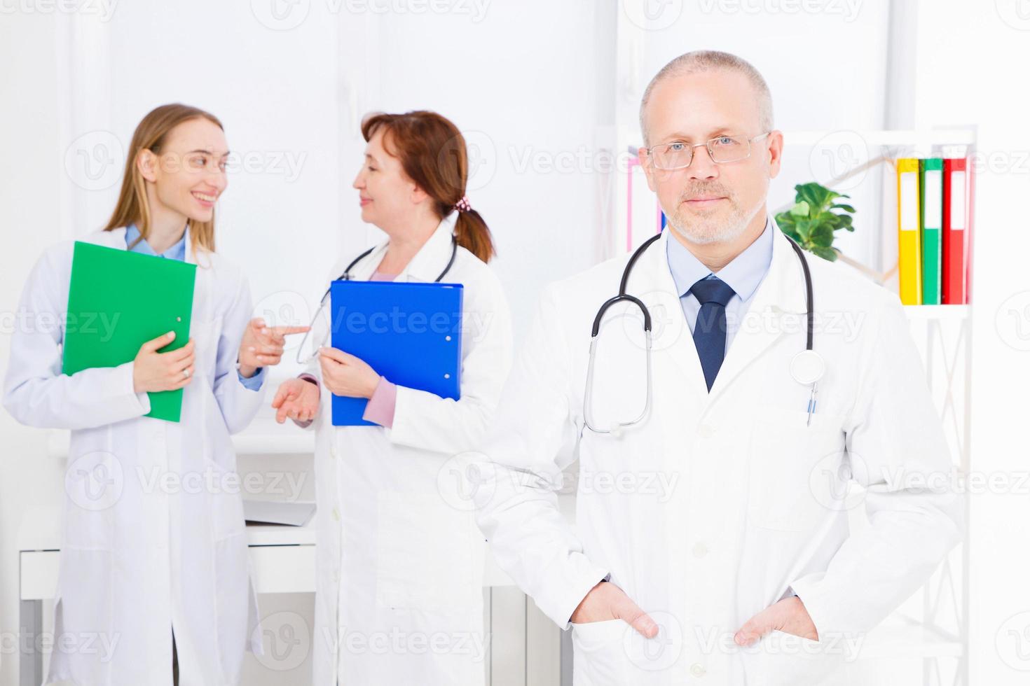 läkare poserar på kontoret, han bär ett stetoskop, medicinsk personal i bakgrunden. kvalitetsmedicin koncept. kopieringsutrymme foto