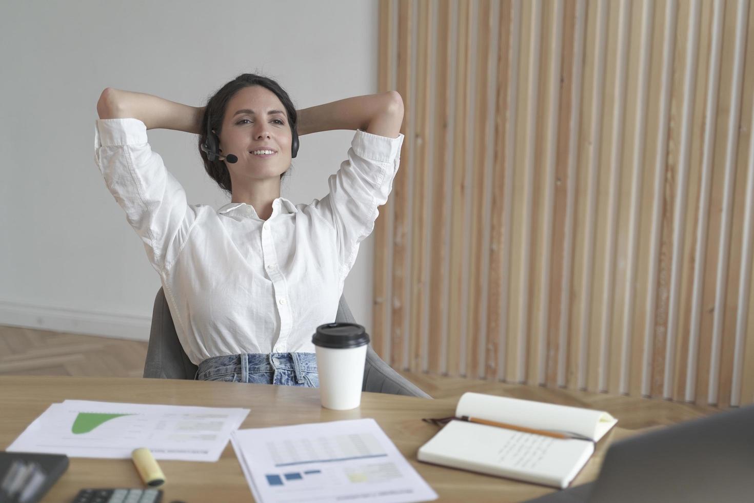 meditativ kvinnlig entreprenör i hörlurar sitter i avslappnad position med händerna bakom huvudet foto