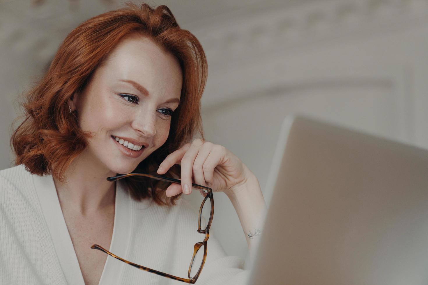 professionell leende ingefära kvinnlig webbdesigner använder applikation på bärbar dator för att skapa projektarbete, gör onlineforskning och surfar på internet, håller glasögon, har ett glatt uttryck foto