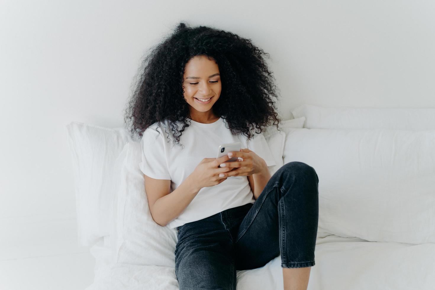 avslappnad vacker afro-kvinna med glada uttryck använder modern mobiltelefon, surfar på internet, poserar i vit bekväm säng, ansluten till wifi, surfar på webbsidan, njuter av inhemsk atmosfär foto