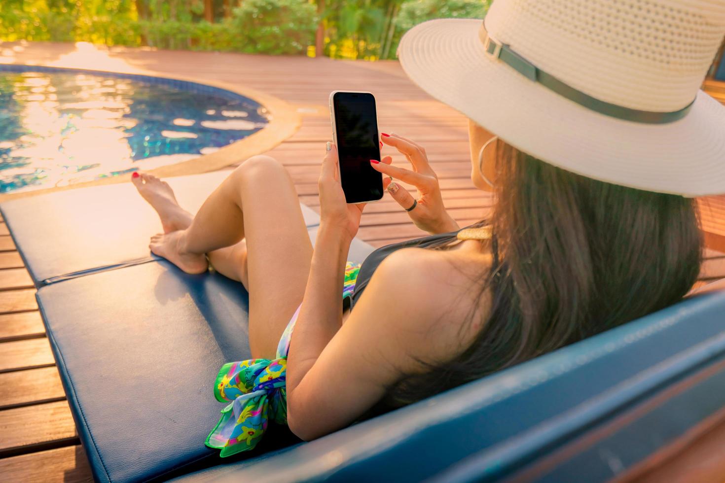 asiatisk kvinna med hatt och baddräkt sitter i stolen vid poolen och använder smartphone på sommarlovet. lyxliv. kollar nyheter på mobiltelefonapplikationen vid poolen. flicka med nagelmanikyr foto