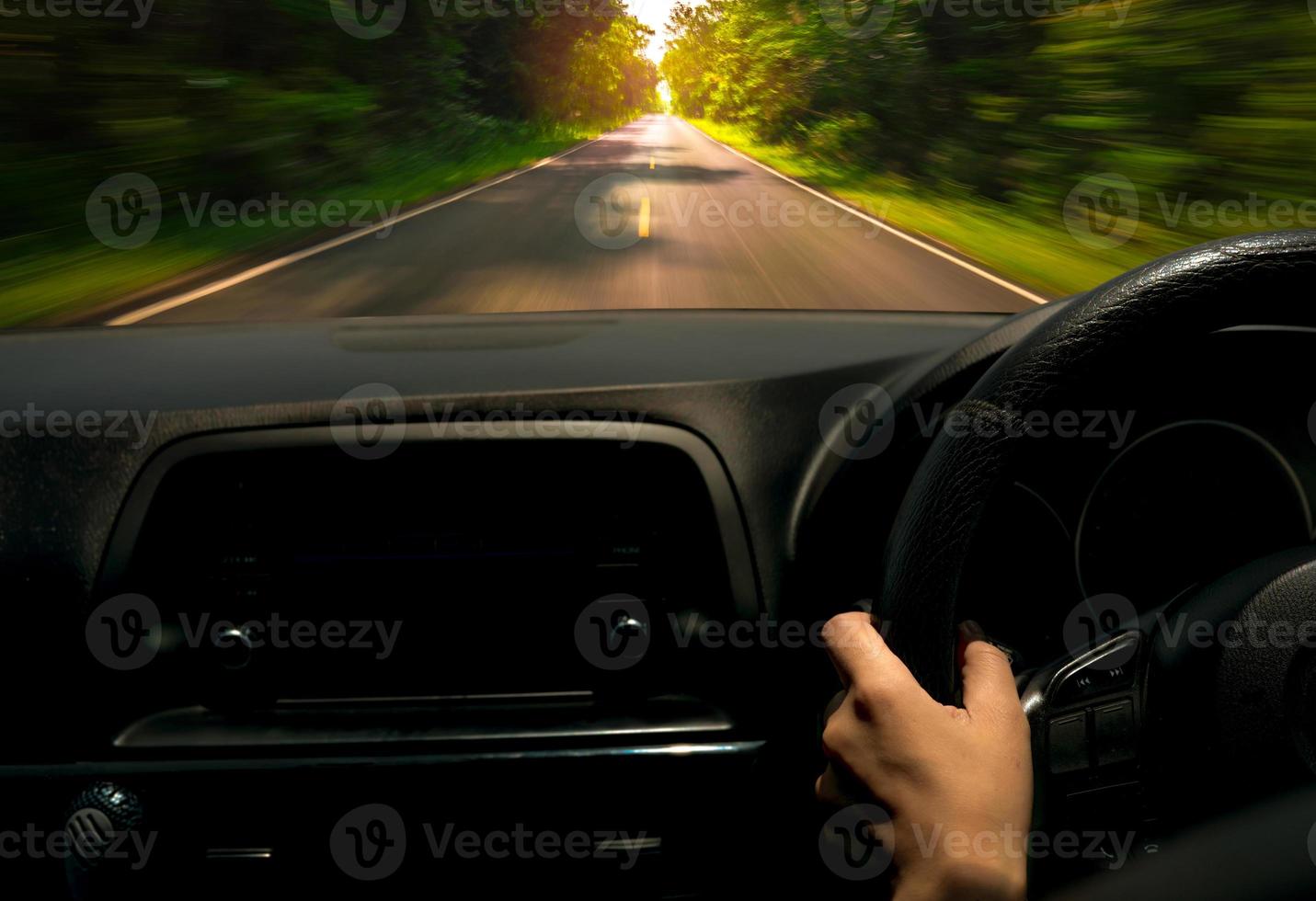 förarhand som håller ratten för kontrollbil. inifrån av bilen. instrumentbräda och vindruta. kör bil på asfaltsväg med rörelseoskärpa i den gröna skogen. perspektivvy snabb bil på motorvägen foto