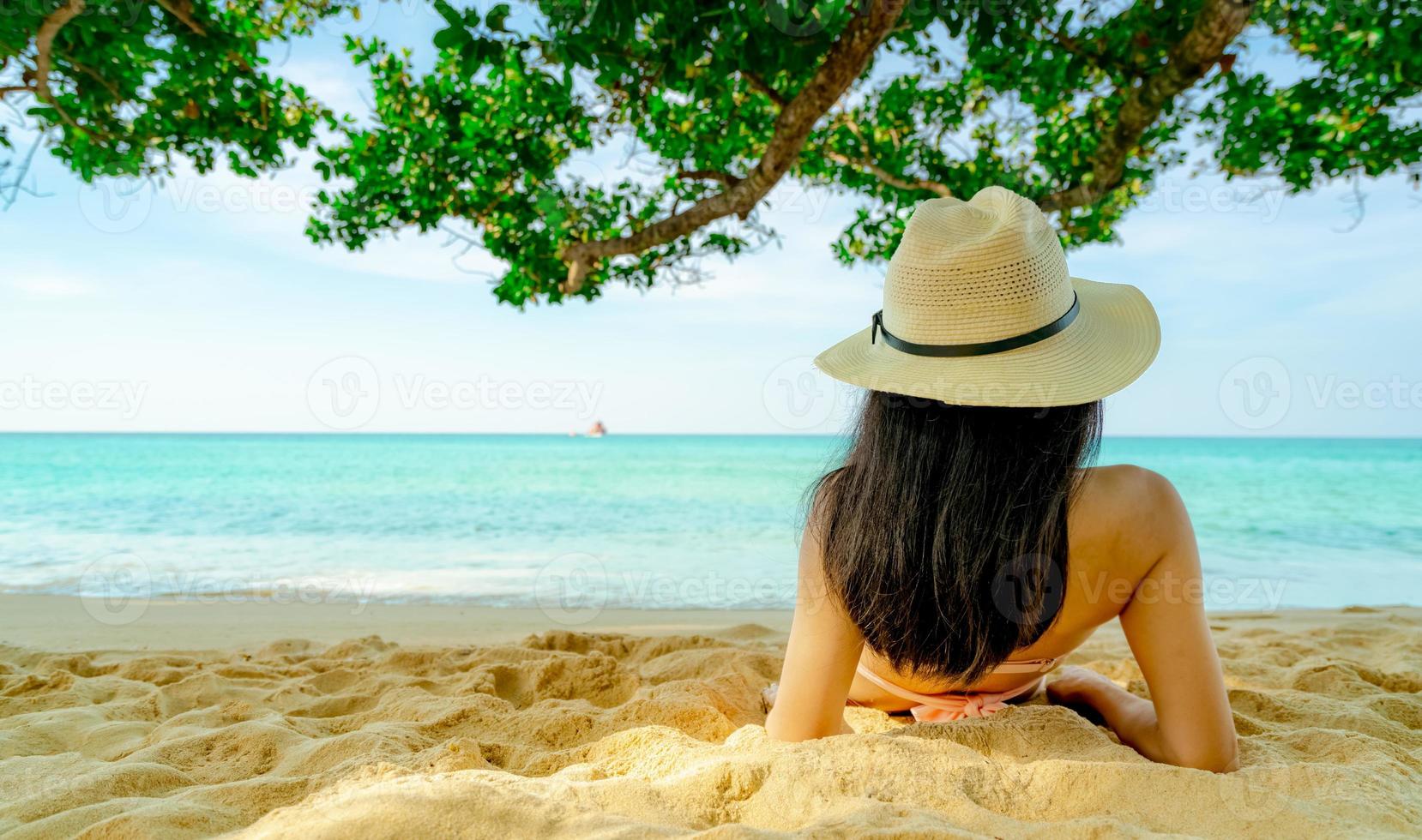 bakifrån av glad ung asiatisk kvinna i rosa baddräkt och halmhatt koppla av och njut av semestern på den tropiska sandstranden under trädet. flicka i sommarlovet mode. skönhet sexig modell. sommarvibbar. foto