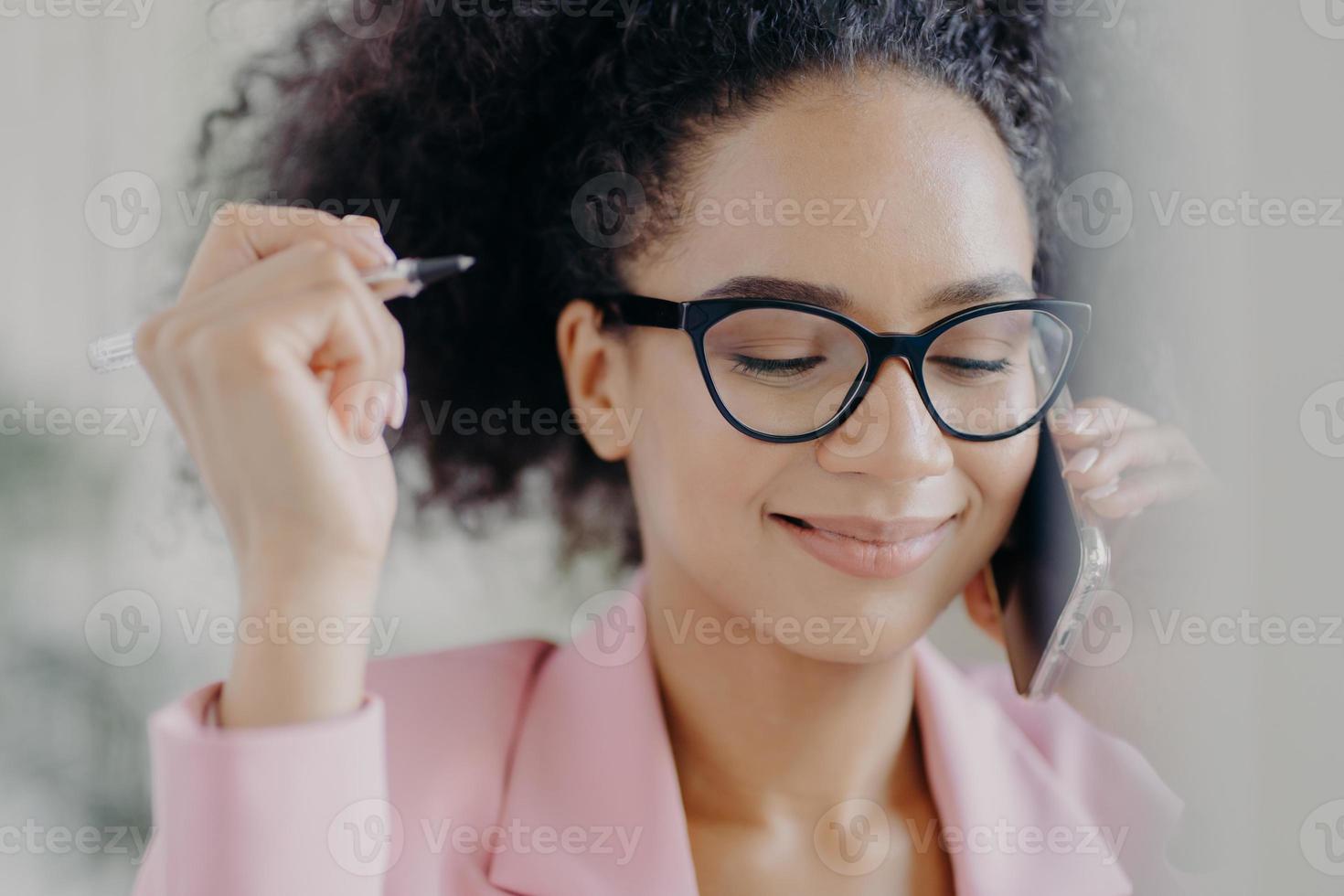 huvudbild av förtjust kvinnlig regissör håller pennan i handen, bär optiska glasögon, har ett milt leende, ringer via modern mobiltelefon, klädd i rosa elegant kostym, fokuserad med blyghet foto