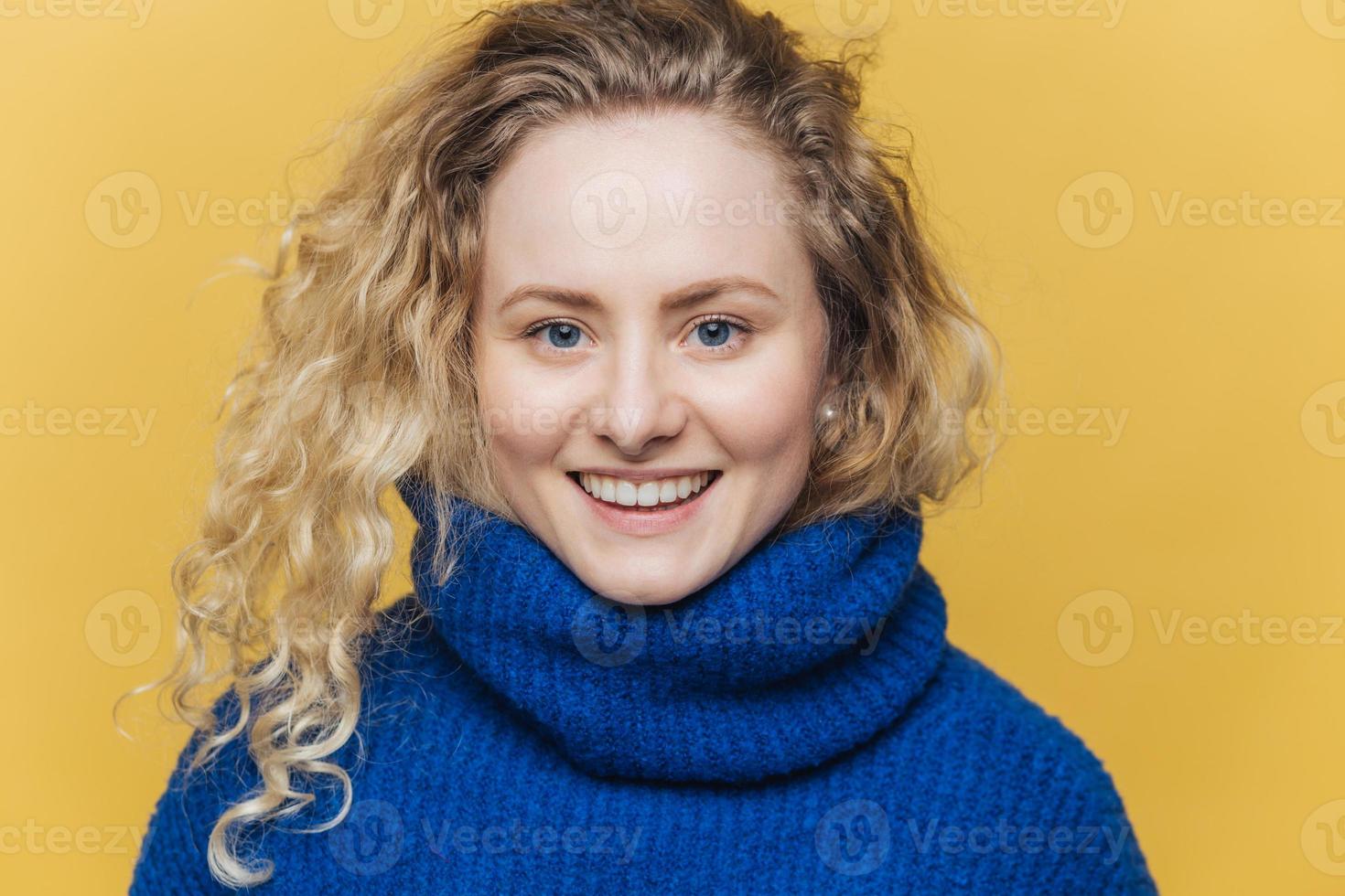 glad glad kvinna med ljust hår, bär varm blå tröja, har ett brett leende och vita tänder som är nöjda efter en lyckad dag, isolerad över gul bakgrund. positiva känslor foto