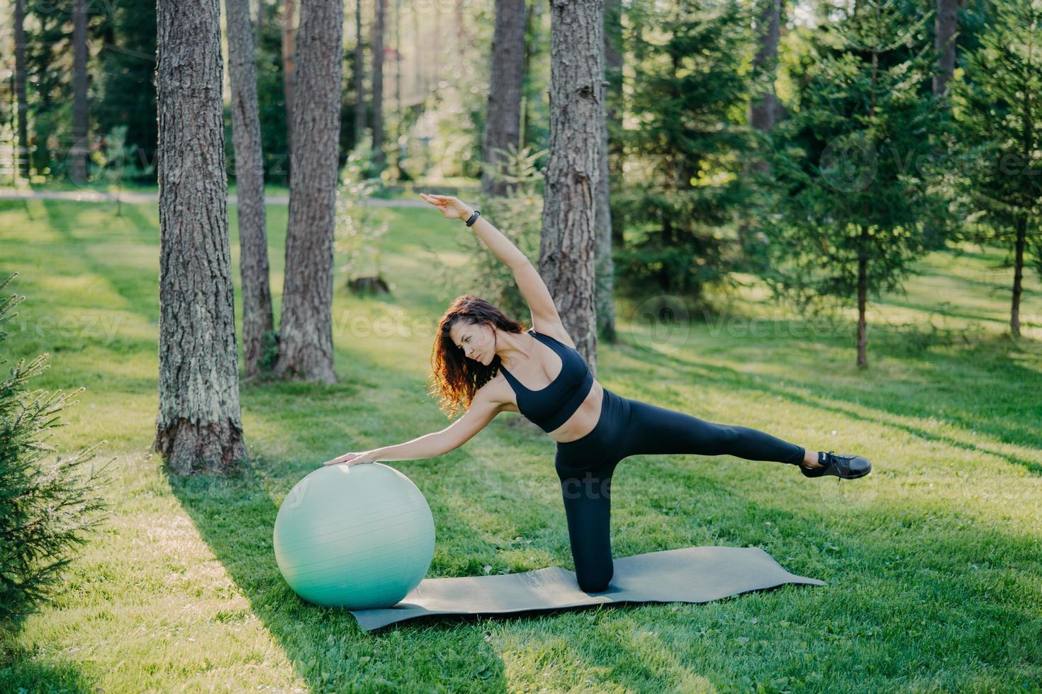 sport och aktiv livsstil koncept. smal sportig kvinna i svarta kläder lutar sig åt sidan på träningsboll, höjer armarna, poserar på karemat i grön park, tränar yoga utomhus, har atletisk kroppsform. foto