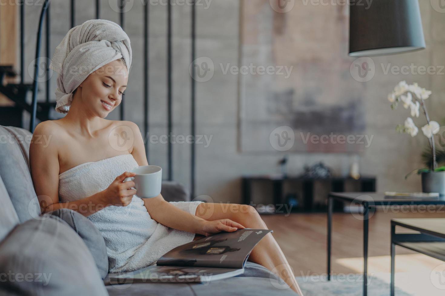 avslappnad leende ung kvinna insvept i handduk efter att ha duschat, dricker kaffe och läser skönhetsmagasin, sitter på en bekväm soffa mot mysig inhemsk interiör. wellness och skönhet koncept foto