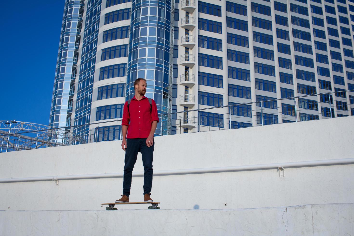 ung skäggig man rider på skateboard, hipster med longboard i röd skjorta och blå jeans urban bakgrund foto