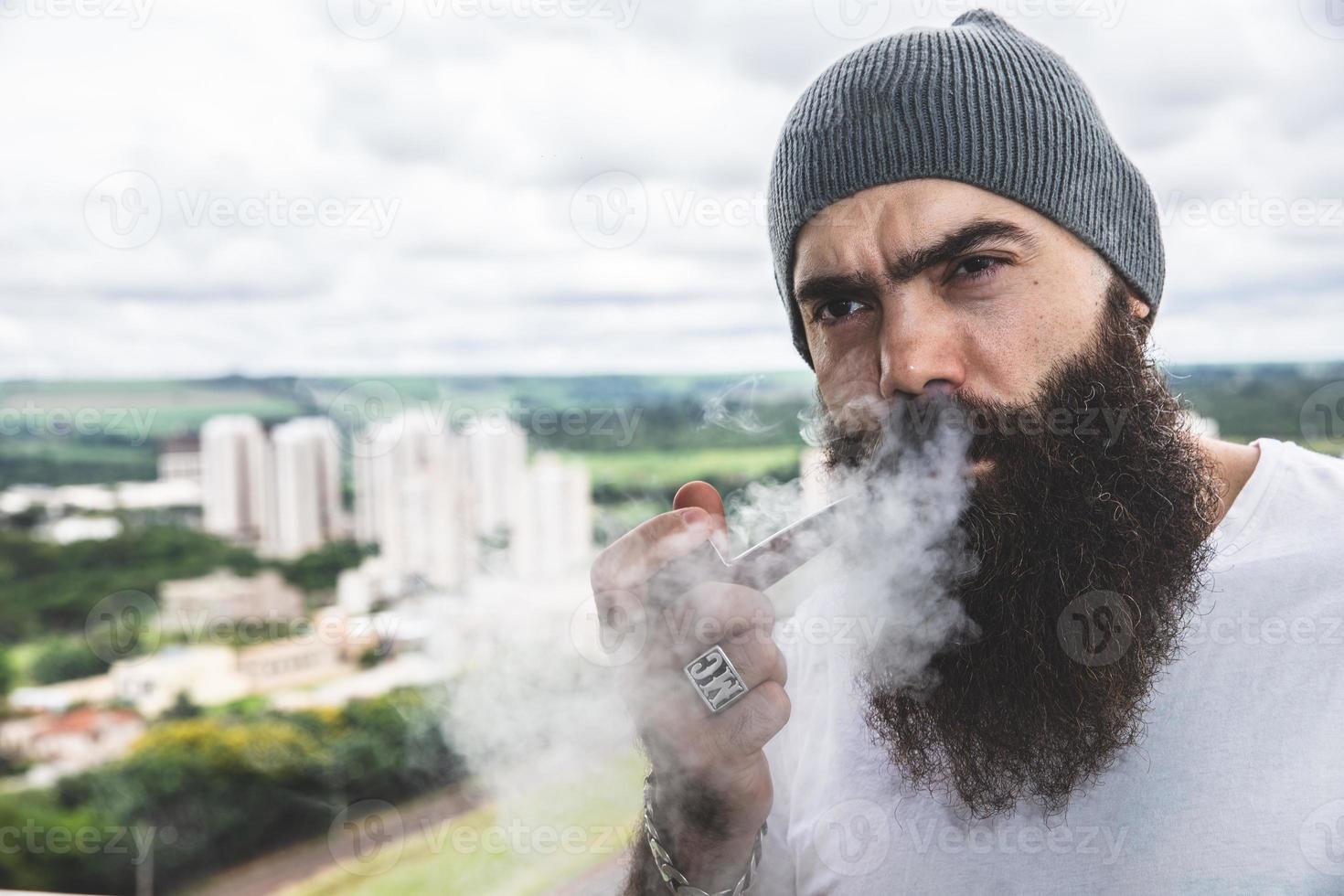 snygg skäggig man röker pipa tittar på horisonten från toppen av en hög byggnad. foto