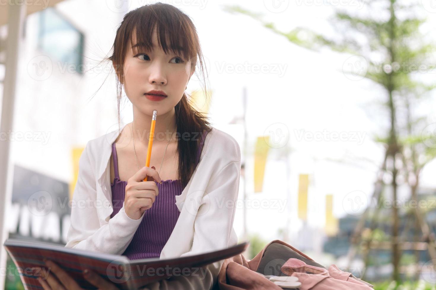 ensamstående ung vuxen asiatisk studentkvinna sitter och studerar med anteckningsboken på campusområdet. foto