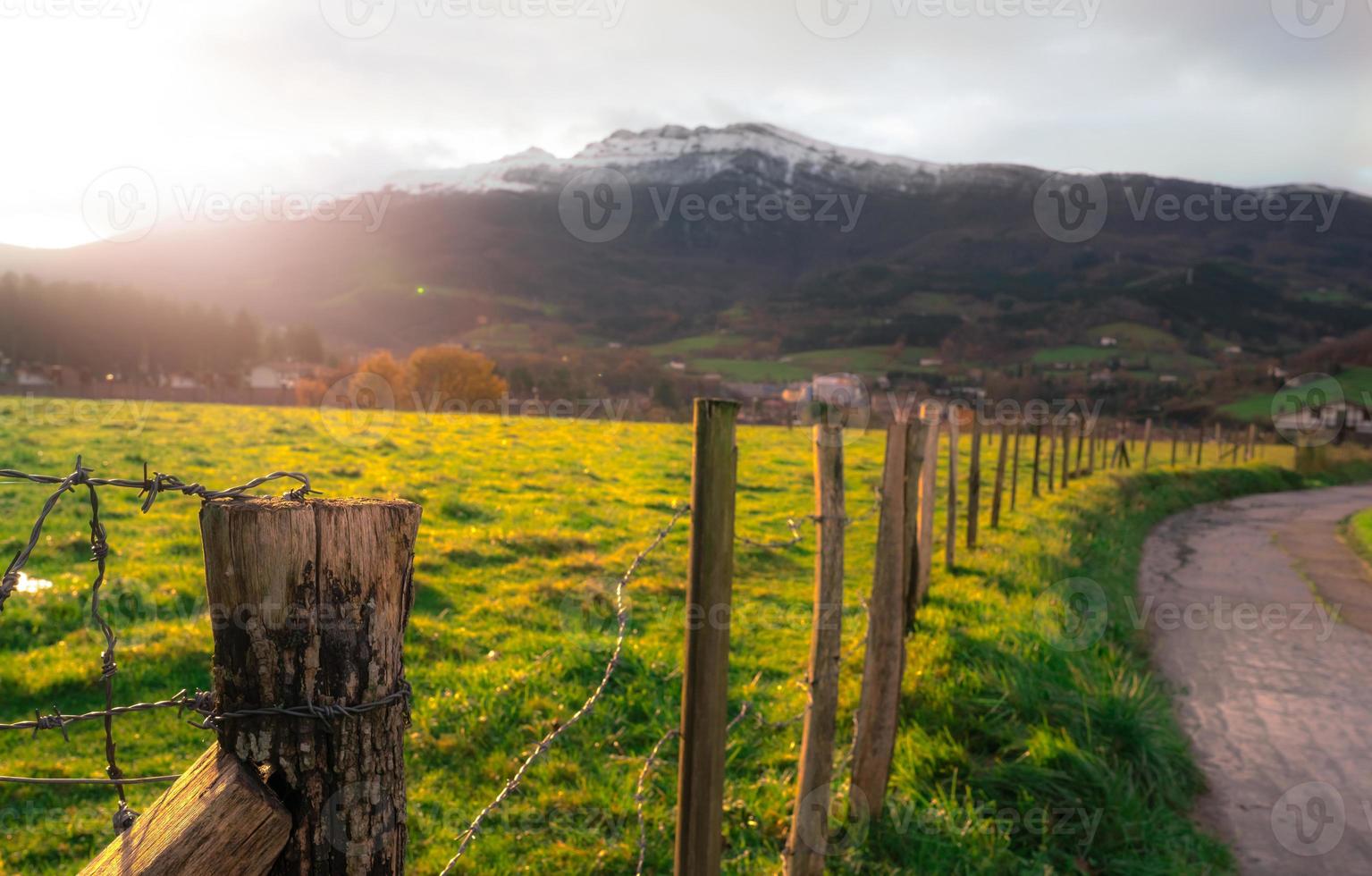 gammal trästolpe av tråd staket på oskärpa grönt gräsfält, stad i dalen och berg i Europa. staket av gröna djur som betar bete bredvid vägen. trästolpe av grönt gräsfält med solljus. foto