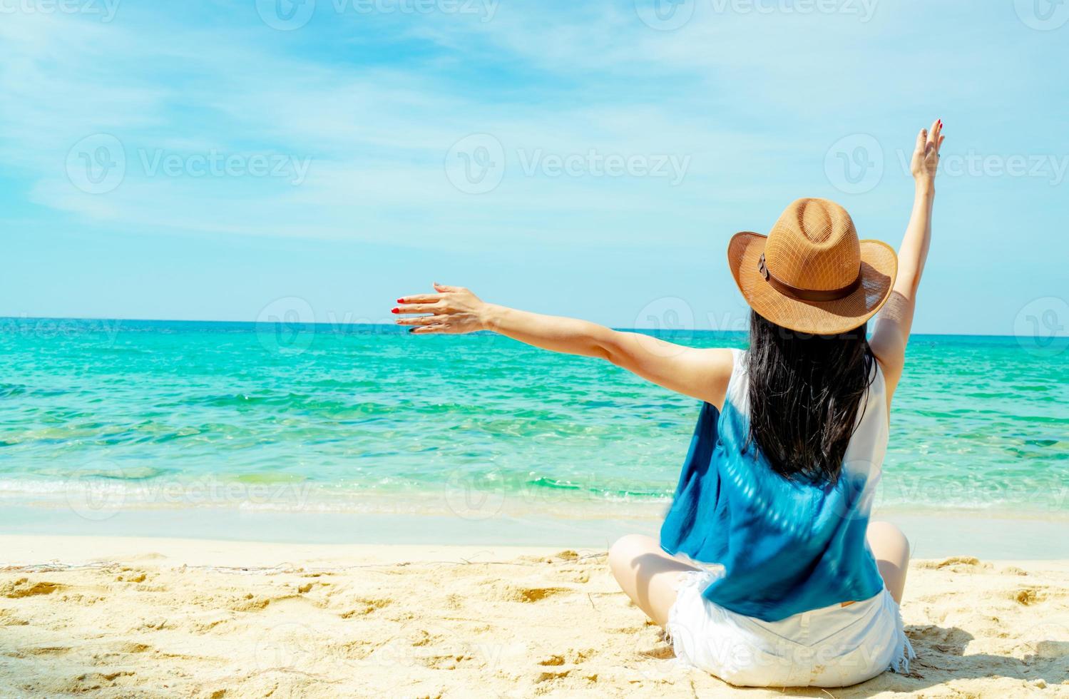 glad ung kvinna bär stråhatt sitta och höjde handen på sandstranden. koppla av och njut av semestern på den tropiska paradisstranden med grönt vatten. flicka i sommarlovet. sommarvibbar. bekymmerslöst koncept. foto