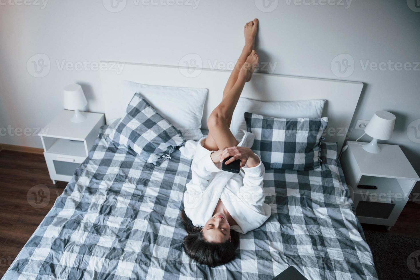 lathetens dag. vacker flicka i vit mantel liggande på sängen på morgonen och använder smartphone foto