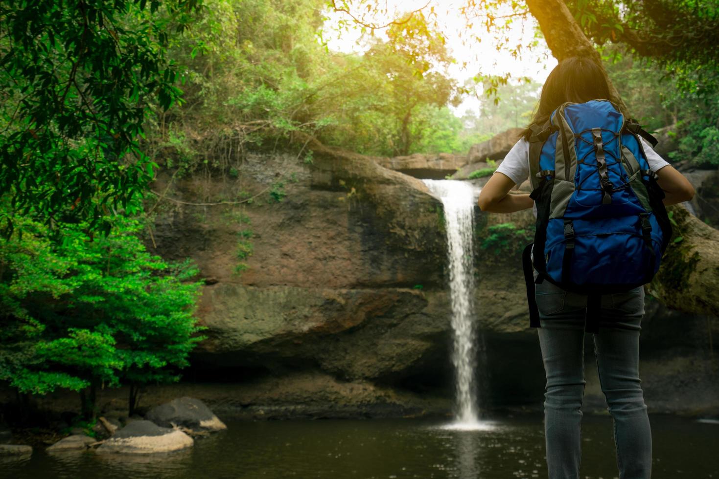 bakifrån av asiatisk kvinna med ryggsäck tittar på litet vattenfall i djungeln. aktiv kvinna med äventyrsresor i grön skog. vandring eller vandring tjej. utomhusaktivitet. vattenfall vid berget. foto