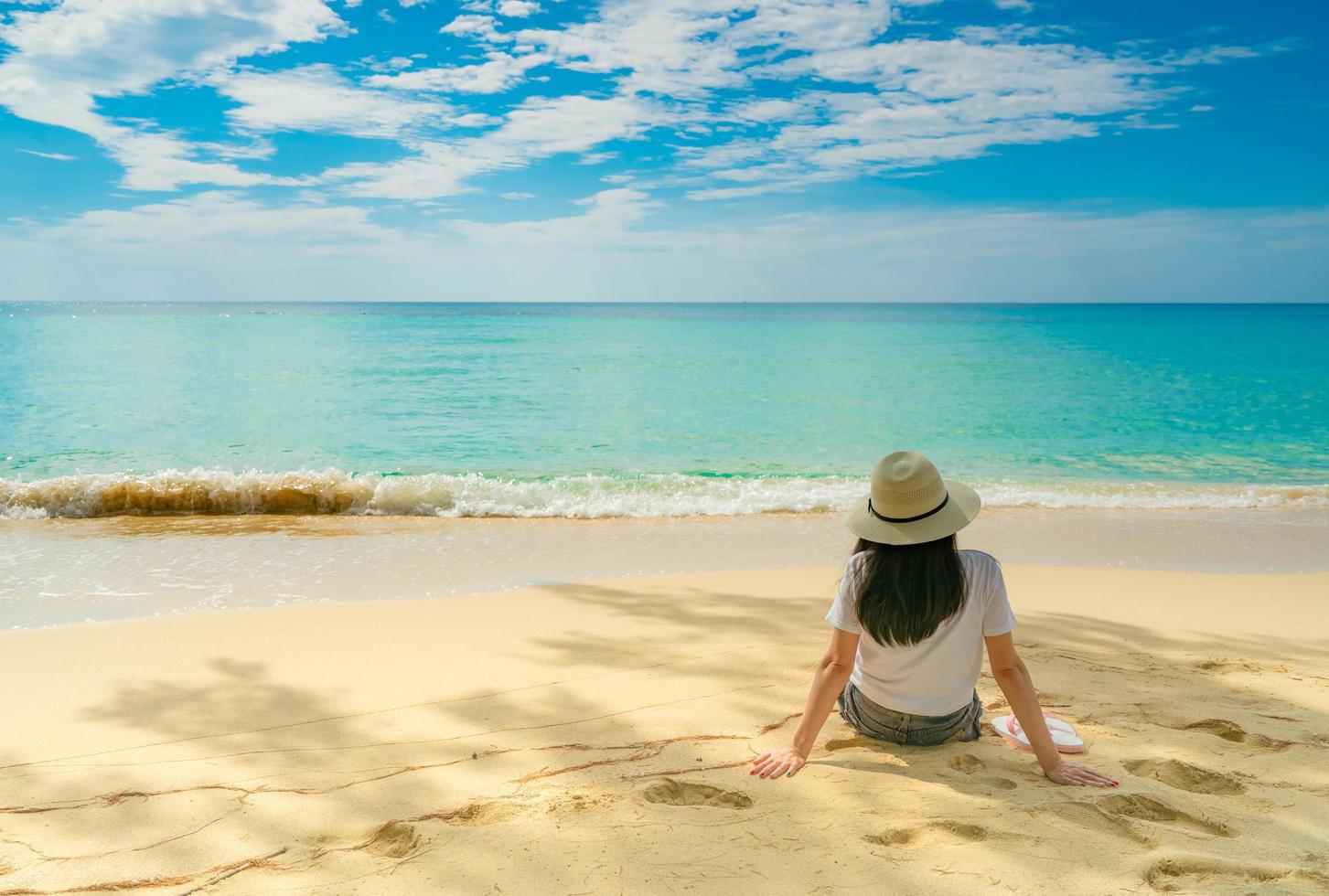 glad ung kvinna i vita skjortor och shorts sitter på sandstranden. koppla av och njuta av semestern på den tropiska paradisstranden med blå himmel och moln. flicka i sommarlovet. sommarvibbar. glad dag. foto