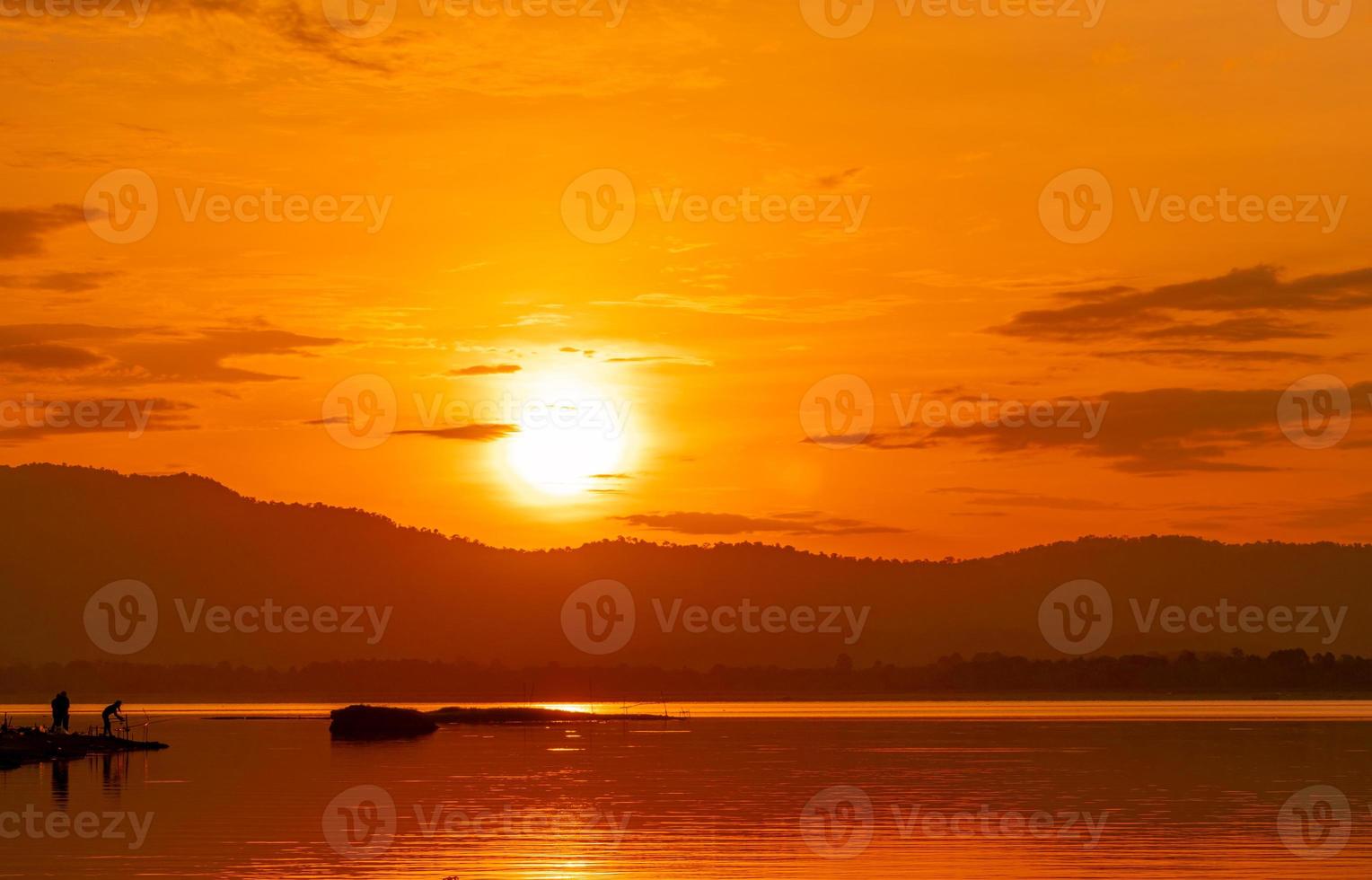 vacker soluppgångshimlen ovanför berget vid reservoaren. människor fiskar med ett fiskespö på floden. landskap av reservoar och berg med orange soluppgång himmel. siluett livet på morgonen. foto
