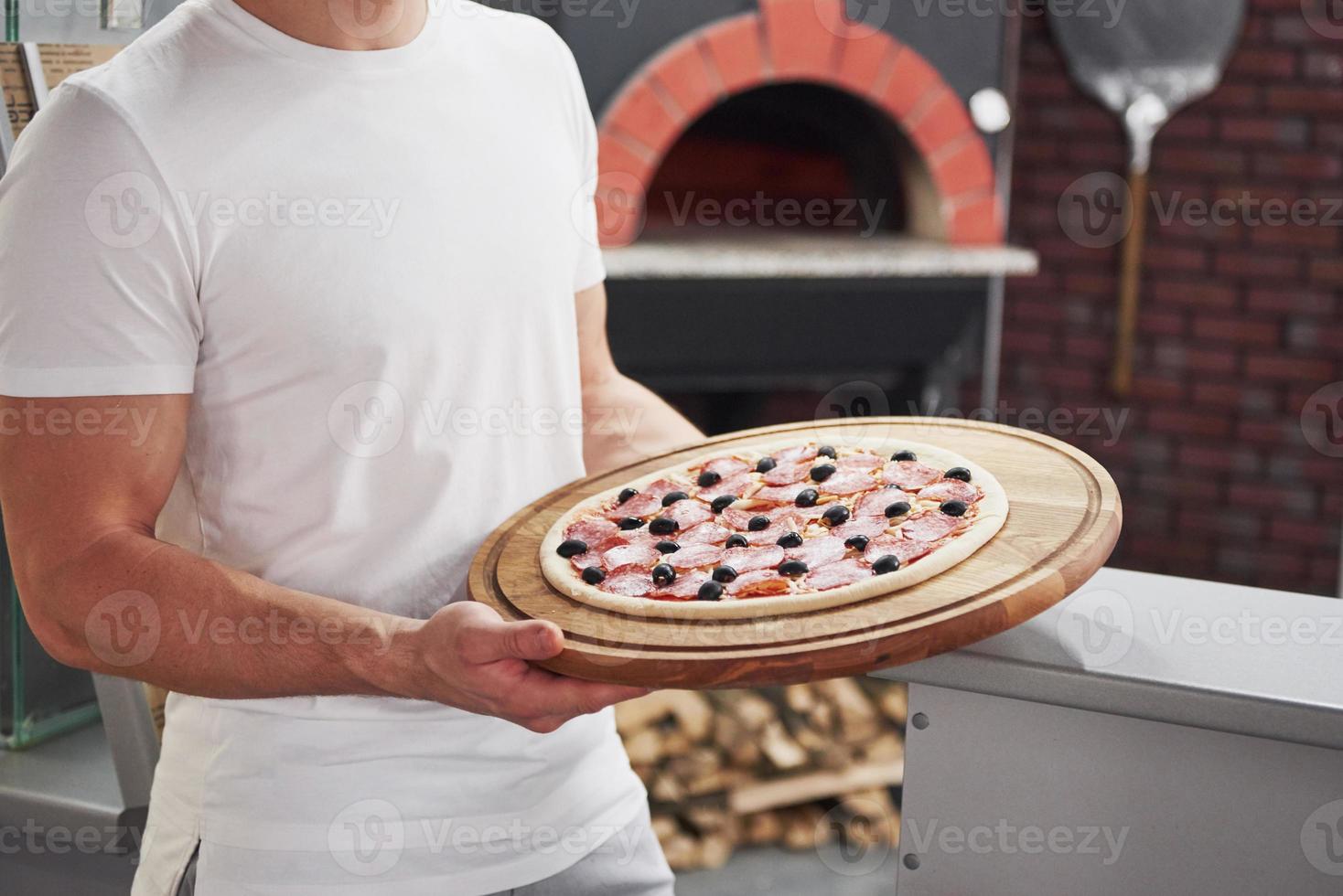 mans händer håller läcker produkt. bagare i vit skjorta med pizza som är redo att sätta i ugnen för att laga mat foto