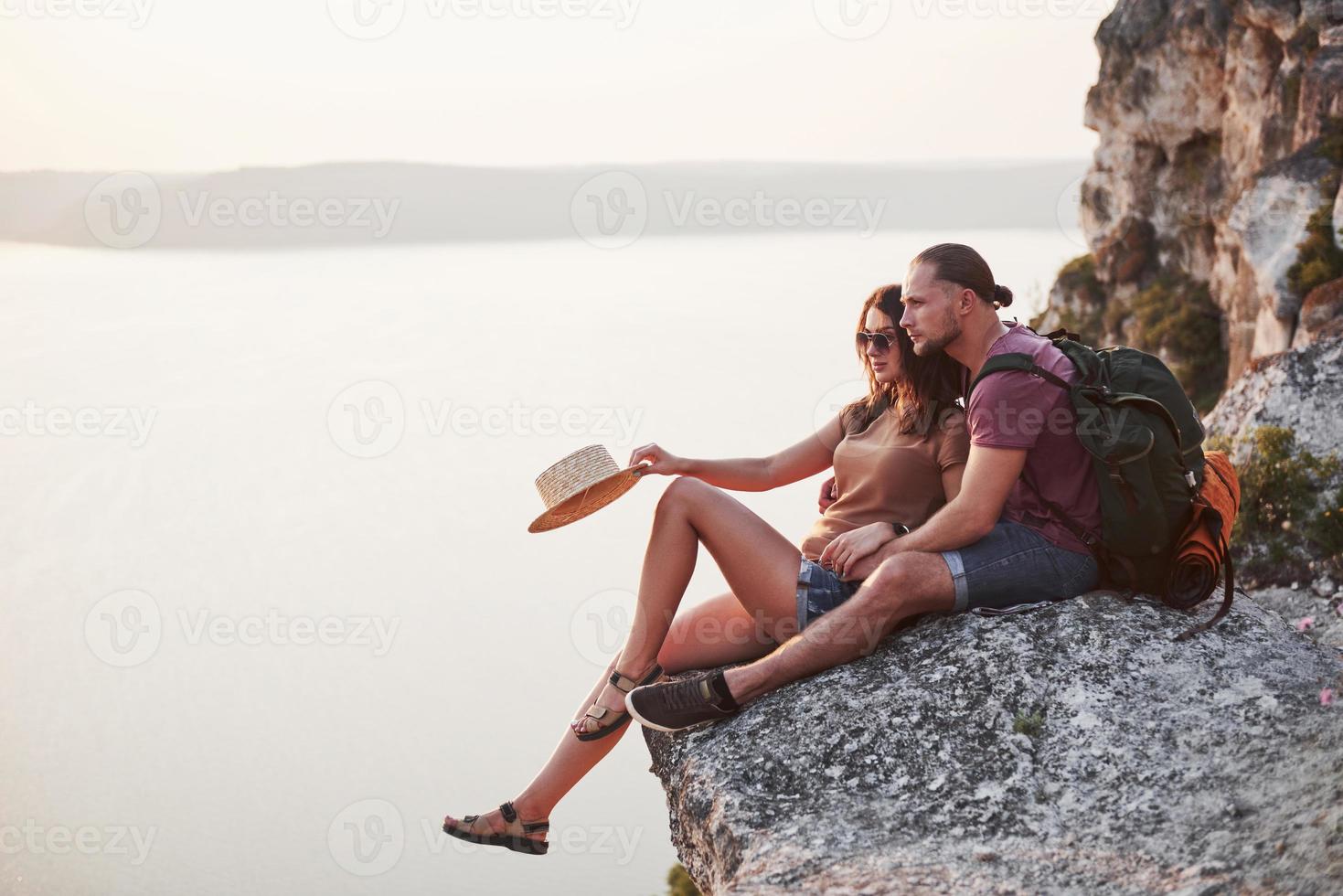 kramar par med ryggsäck sitter på toppen av berget njuter av utsikten kusten en flod eller sjö. resa längs berg och kust, frihet och aktivt livsstilskoncept foto