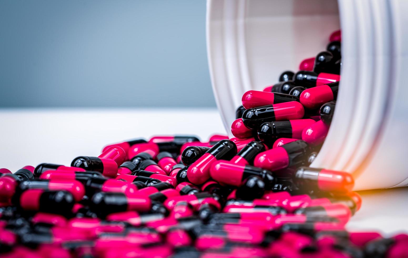 rosa, svarta kapslar piller spills ut från vit plastflaska behållare. receptbelagt läkemedel. antibiotikaresistens. antimikrobiella kapselpiller. läkemedelsindustri. apotek bakgrund. foto
